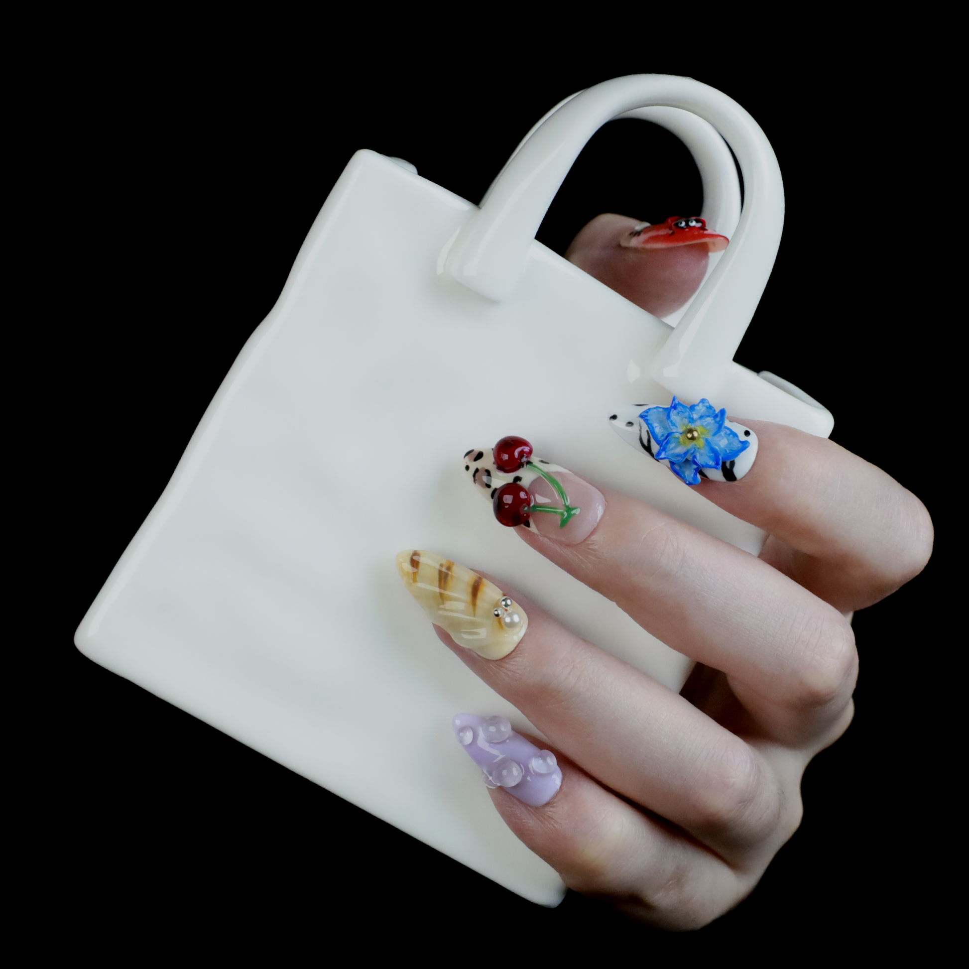 The Garden Blooms Press-On Nails by We Do Nails feature a whimsical garden theme with long, decorative nails, including a blue flower, cherries, red and white patterns, pearls, and stripes. Set against a black background, the hand is partially covered by a black sleeve.