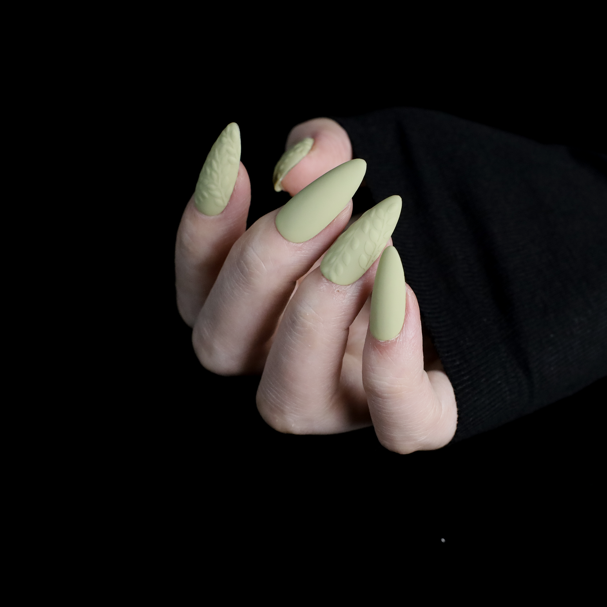 A person displays their crossed hands against a black background, showcasing We Do Nails Earthy Olive Green Press-On Nails. The textured olive design is elegantly highlighted by the sleeves of their black garment.