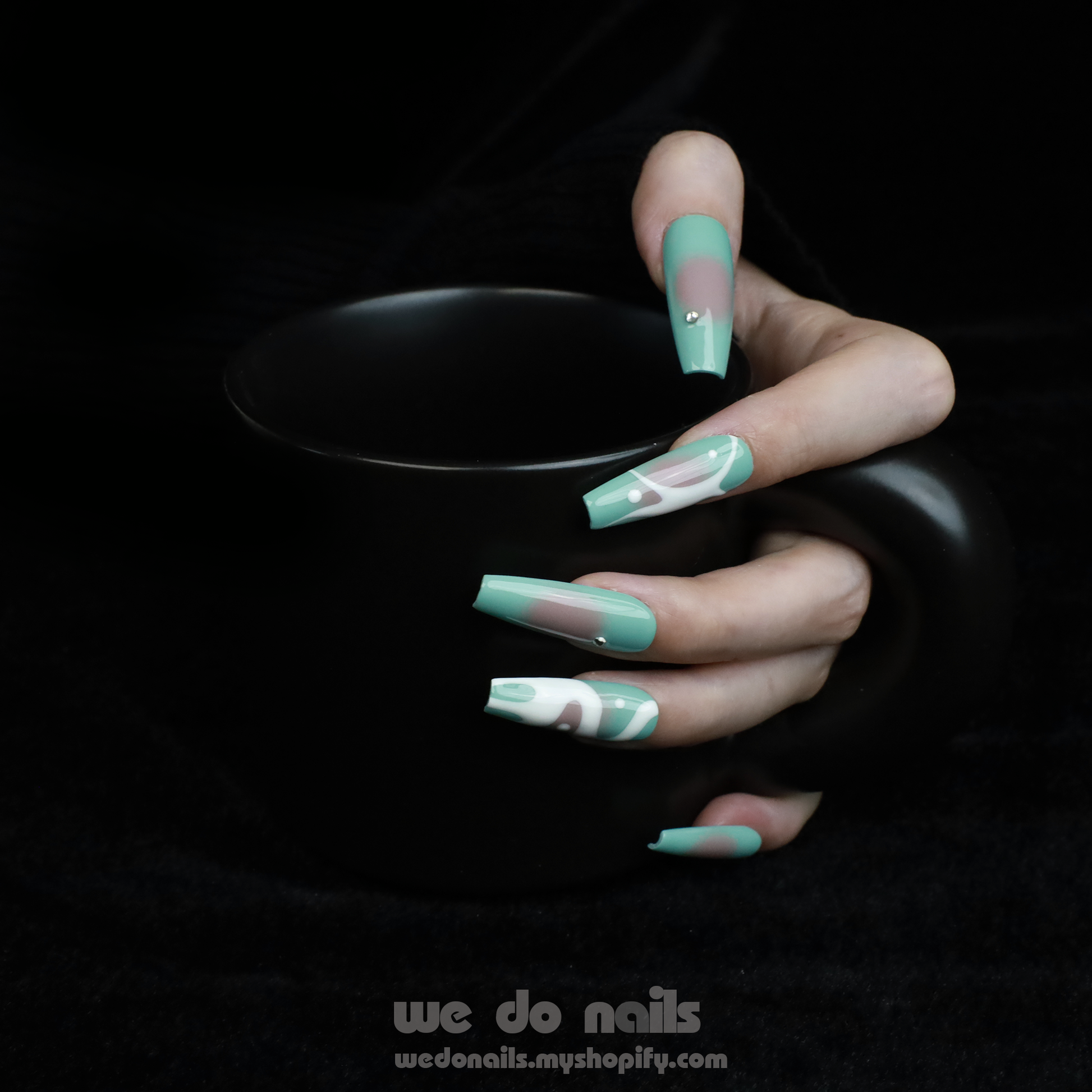Hands with long, angular green nails pose gracefully against a black background, showcasing a modern and stylish design. The glossy finish and rings add flair, elegantly highlighting the allure of Spilled Milk Press-On Nails by We Do Nails.