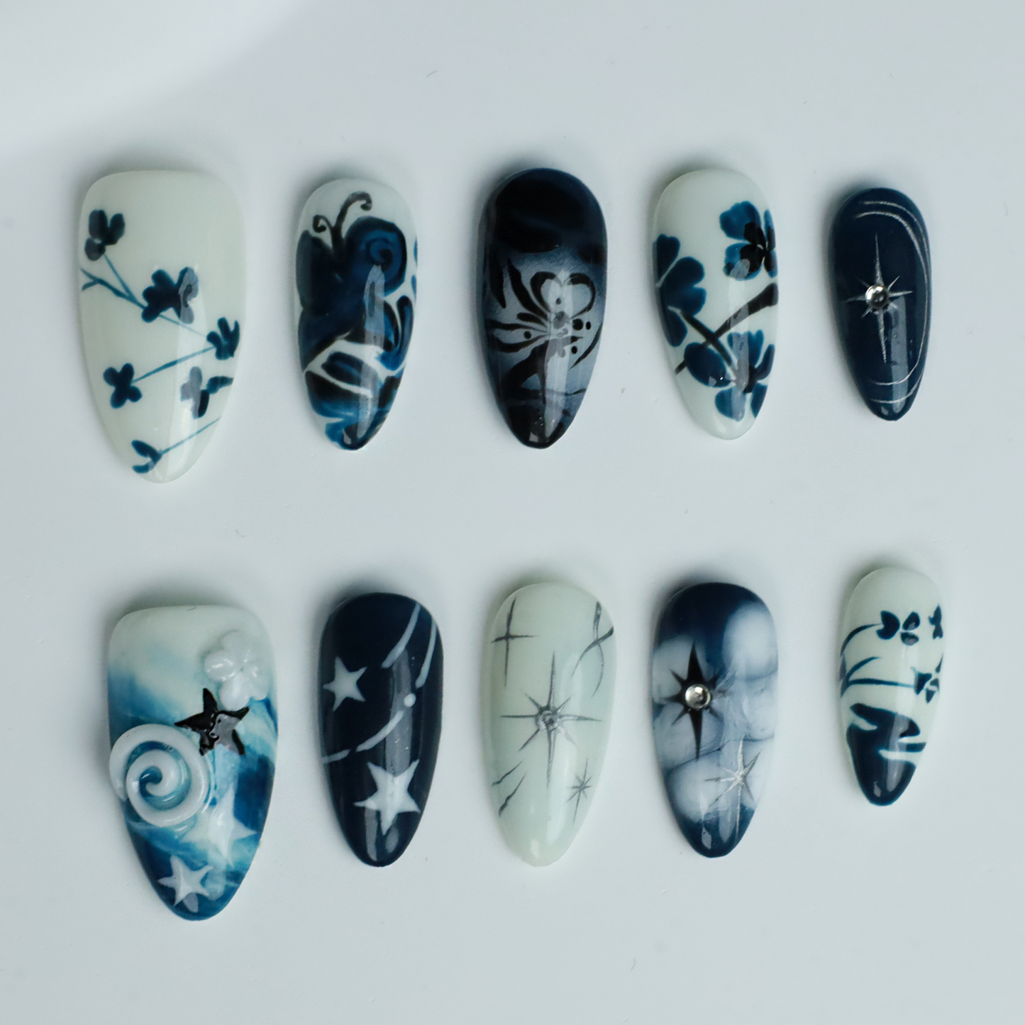 Hands adorned with We Do Nails Blue & White Porcelain Inspired Nails, showcasing long, almond-shaped designs with intricate traditional floral and abstract patterns. The nails stand out against black sleeves and a black background, reflecting the elegance of Chinese porcelain.