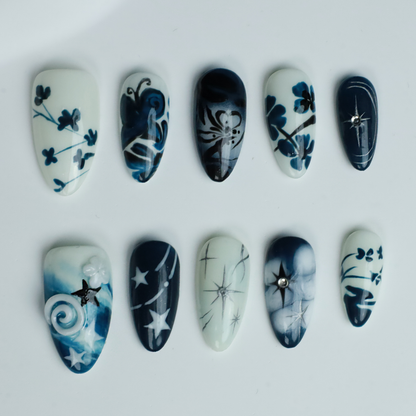 Hands adorned with We Do Nails Blue & White Porcelain Inspired Nails, showcasing long, almond-shaped designs with intricate traditional floral and abstract patterns. The nails stand out against black sleeves and a black background, reflecting the elegance of Chinese porcelain.