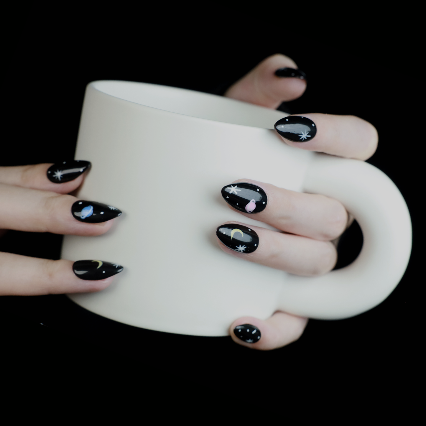 Hands decorated with Galactic Night Sky Press On Nails by We Do Nails, featuring stars, moons, and planets, hold a plain white mug against a dark backdrop.