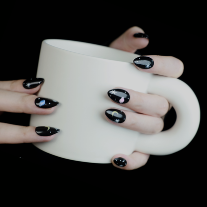 Hands decorated with Galactic Night Sky Press On Nails by We Do Nails, featuring stars, moons, and planets, hold a plain white mug against a dark backdrop.