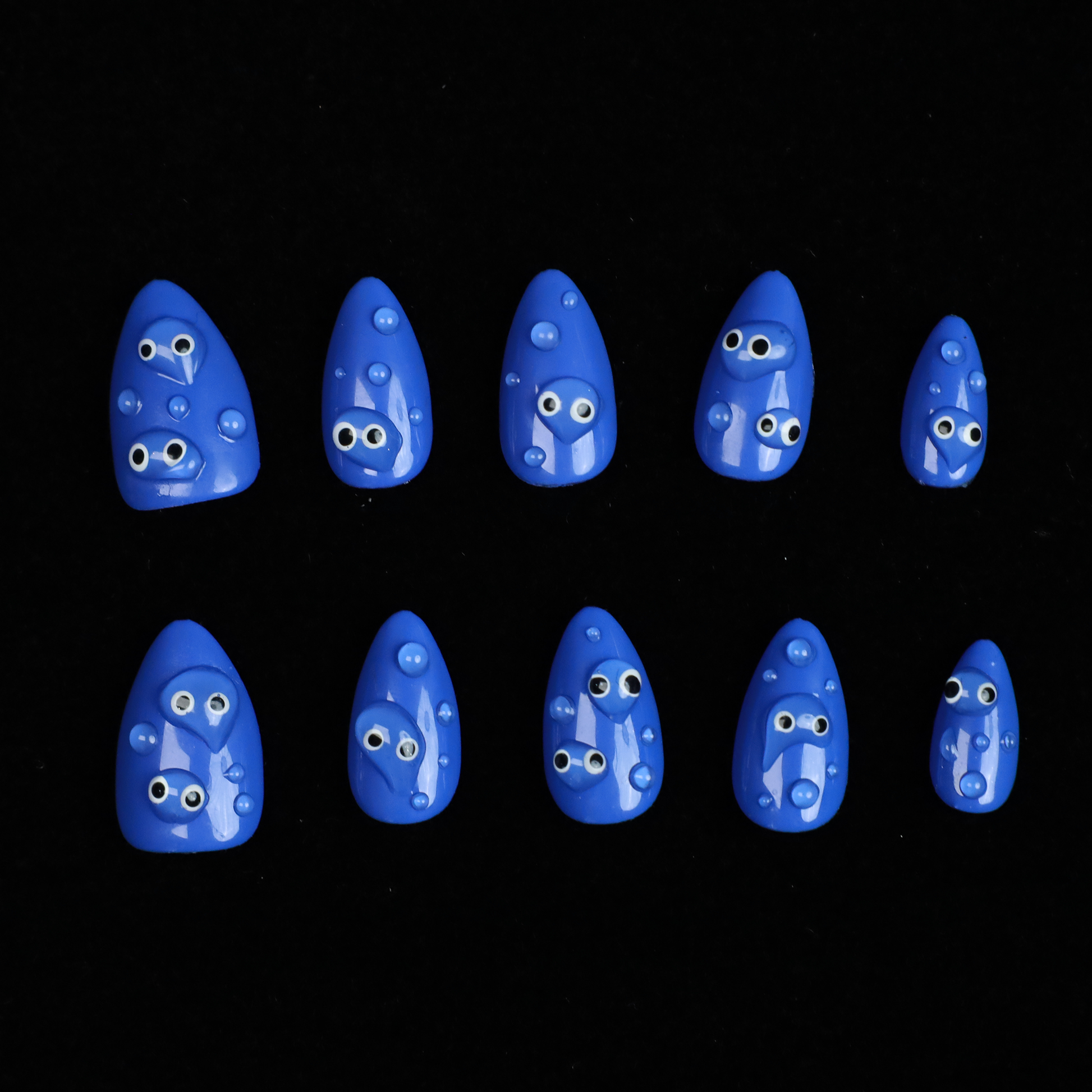 A hand with We Do Nails Cute Blue Monster Press-On Nails set features long, almond-shaped, bright blue nails adorned with playful cartoon faces and 3D googly eyes. The hand in a black sweater is striking against the dark background.