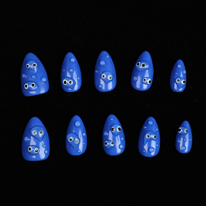 A hand with We Do Nails Cute Blue Monster Press-On Nails set features long, almond-shaped, bright blue nails adorned with playful cartoon faces and 3D googly eyes. The hand in a black sweater is striking against the dark background.