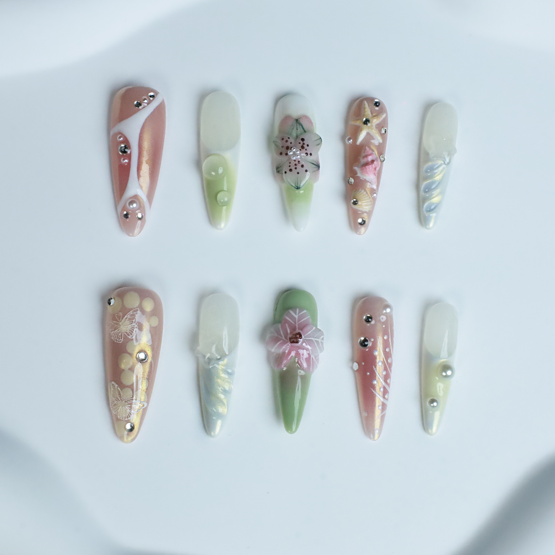 A hand flaunts We Do Nails Floral Blush Pearl Press-On Nails: green with pearls, pink with a butterfly, clear with gold accents, blue with a wave pattern, and a pink-white thumb—all set against a black background for an enchanting look.