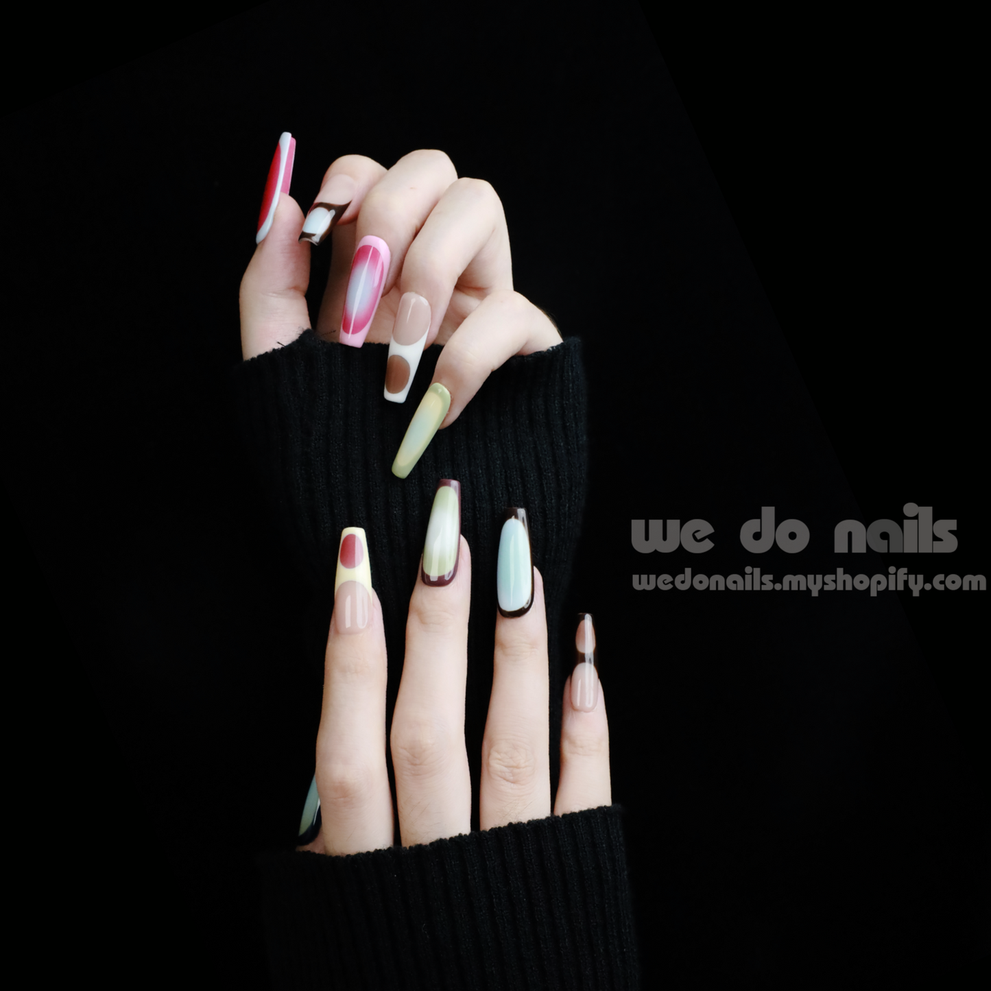 A pair of hands with long, artistic Ms.Circles Press-On Nails by We Do Nails stand out against a black background. The vibrant nails have bold patterns in green, pink, and nude hues. Partially emerging from black sweater sleeves, they create a striking contrast.