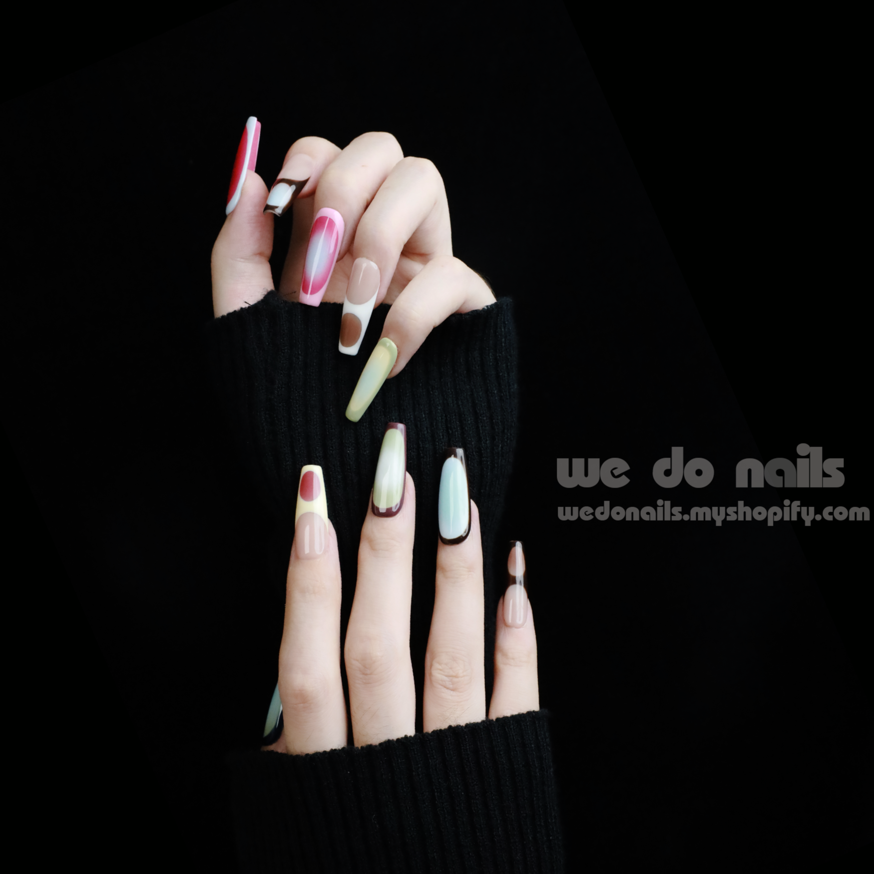 A pair of hands with long, artistic Ms.Circles Press-On Nails by We Do Nails stand out against a black background. The vibrant nails have bold patterns in green, pink, and nude hues. Partially emerging from black sweater sleeves, they create a striking contrast.