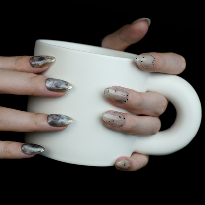 Hands showcasing We Do Nails Marble and Speckled Press On Nails, almond-shaped with a high-gloss shine and scattered speckles, perfectly highlighted against a solid black background.