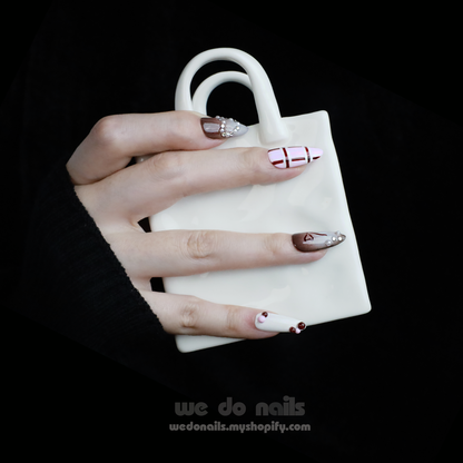 A hand with enchanting nails holds a small white handbag. The Charming Cocoa Delight Press-On Nails from We Do Nails showcase intricate patterns, featuring stripes and dotted embellishments in white, brown, and red shades, all set against a black background that enhances their allure.
