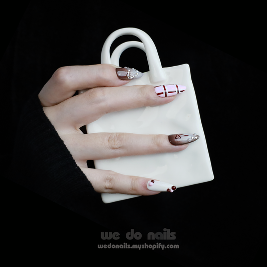A hand with enchanting nails holds a small white handbag. The Charming Cocoa Delight Press-On Nails from We Do Nails showcase intricate patterns, featuring stripes and dotted embellishments in white, brown, and red shades, all set against a black background that enhances their allure.