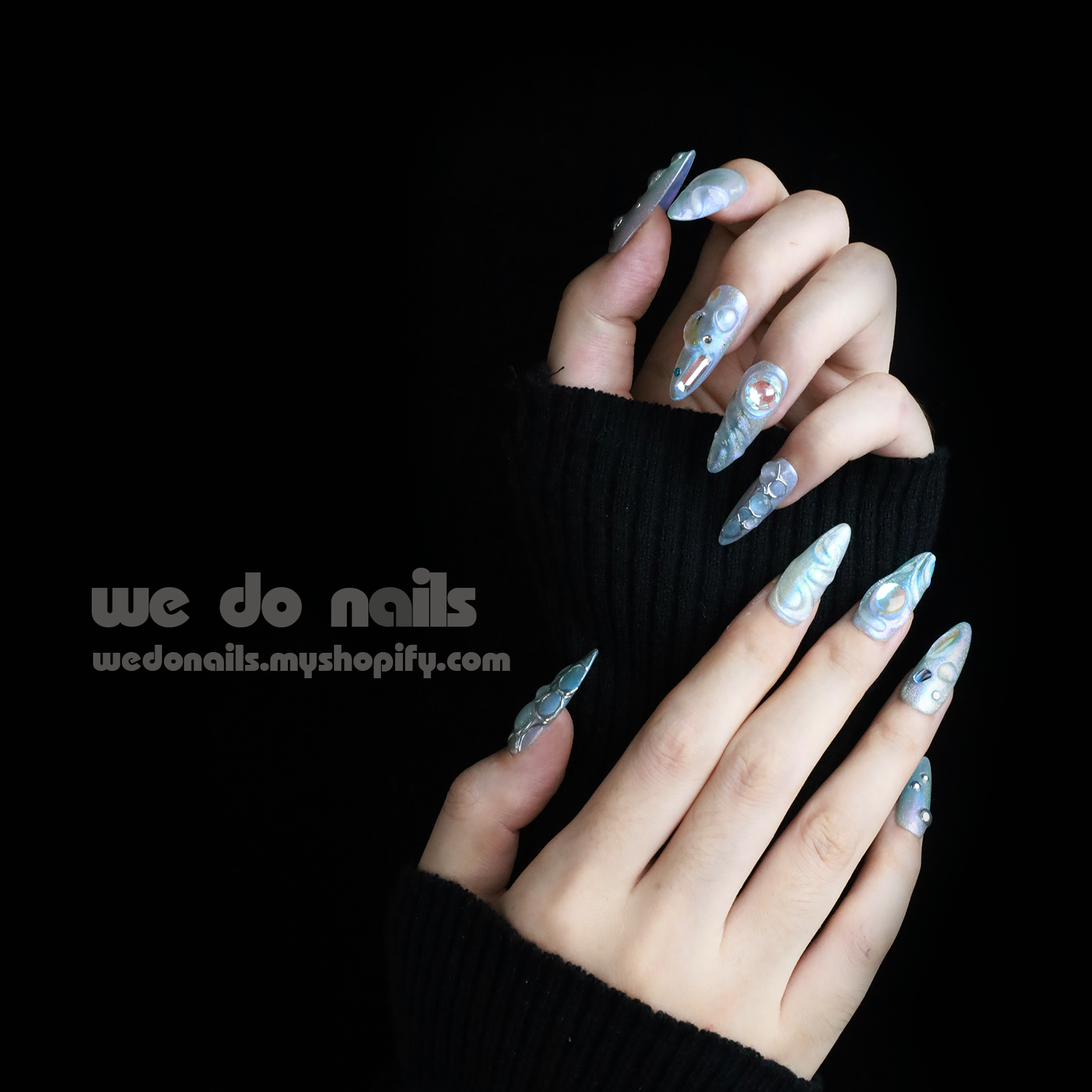 Displaying interlocked fingers adorned with We Do Nails Iridescent Bubble Press-On Nails, these long, pointed artificial nails feature a holographic silver and pearl finish, beautifully contrasted by a sleek black garment. Text reads we do nails and wedonails.myshopify.com.