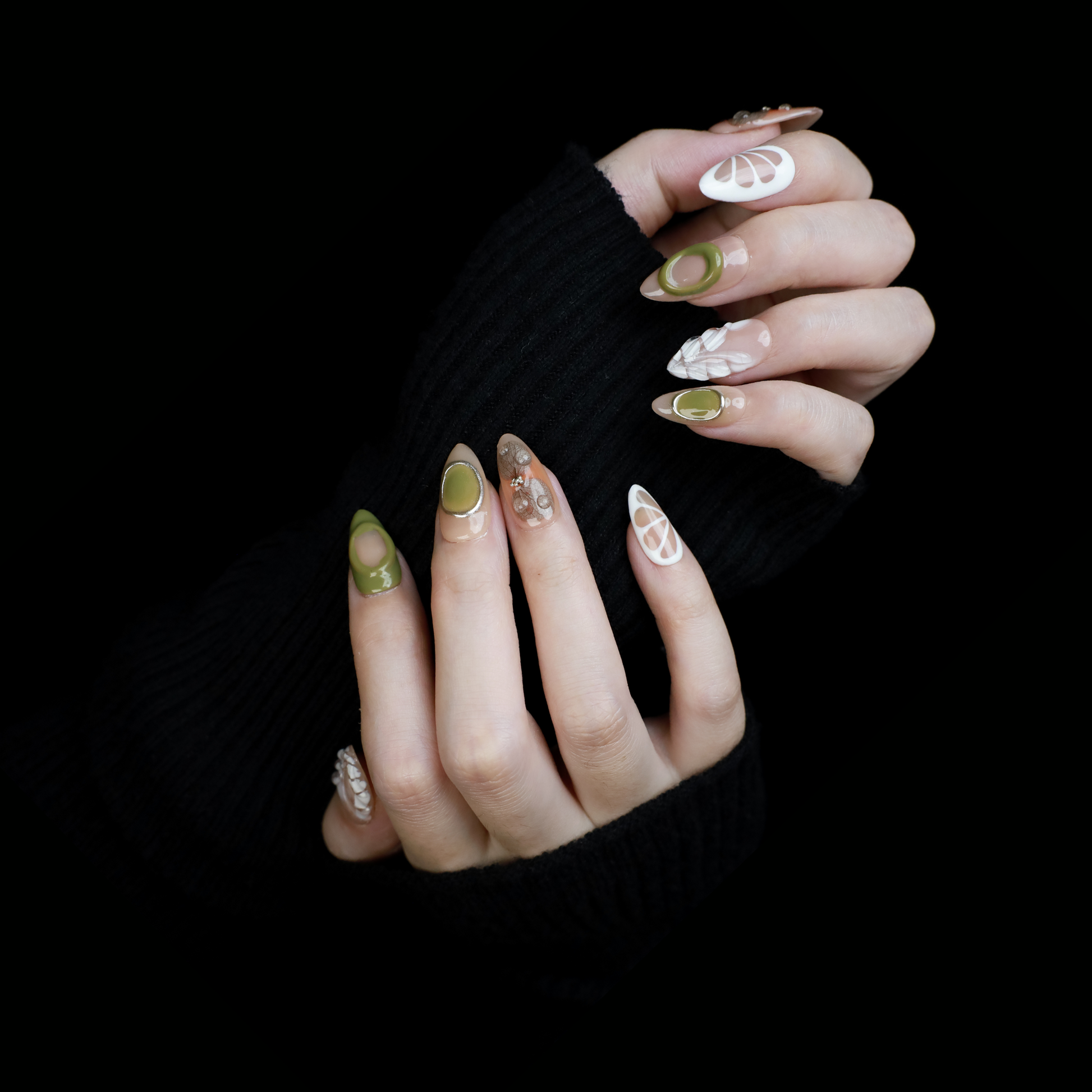 A hand in a black sleeve displays the Leaf and Organic design Press-On Nails by We Do Nails, featuring long acrylic nails with white abstract patterns, green accents, and clear 3D embellishments that create a nature-inspired look against the dark background.