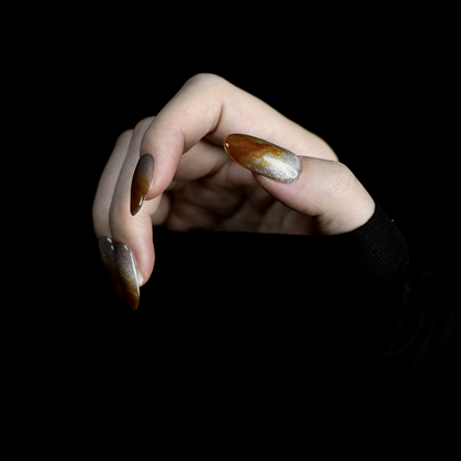 A hand with We Do Nails Elegant Brown Marble Press-On Nails, featuring a subtle metallic chrome accent, holds a simple white mug against a black backdrop.