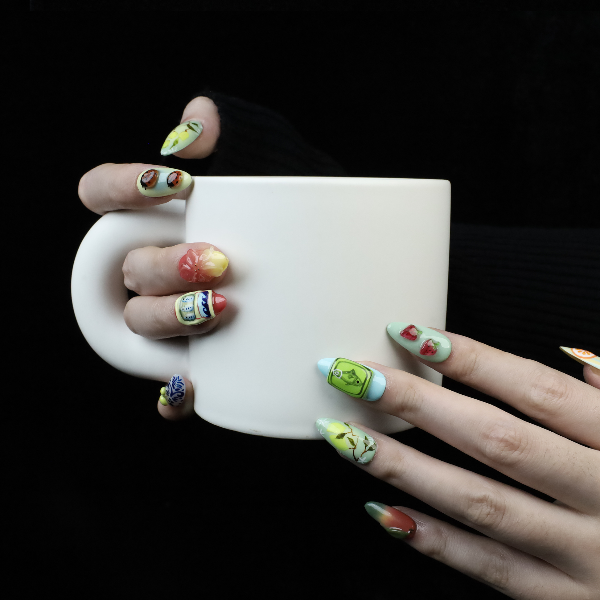 A hand holds a white mug against a black backdrop, flaunting We Do Nails Cute Fruity Vibe Press-On Nails with detailed, colorful designs of strawberries, vegetables, and intricate patterns for a vibrant display.