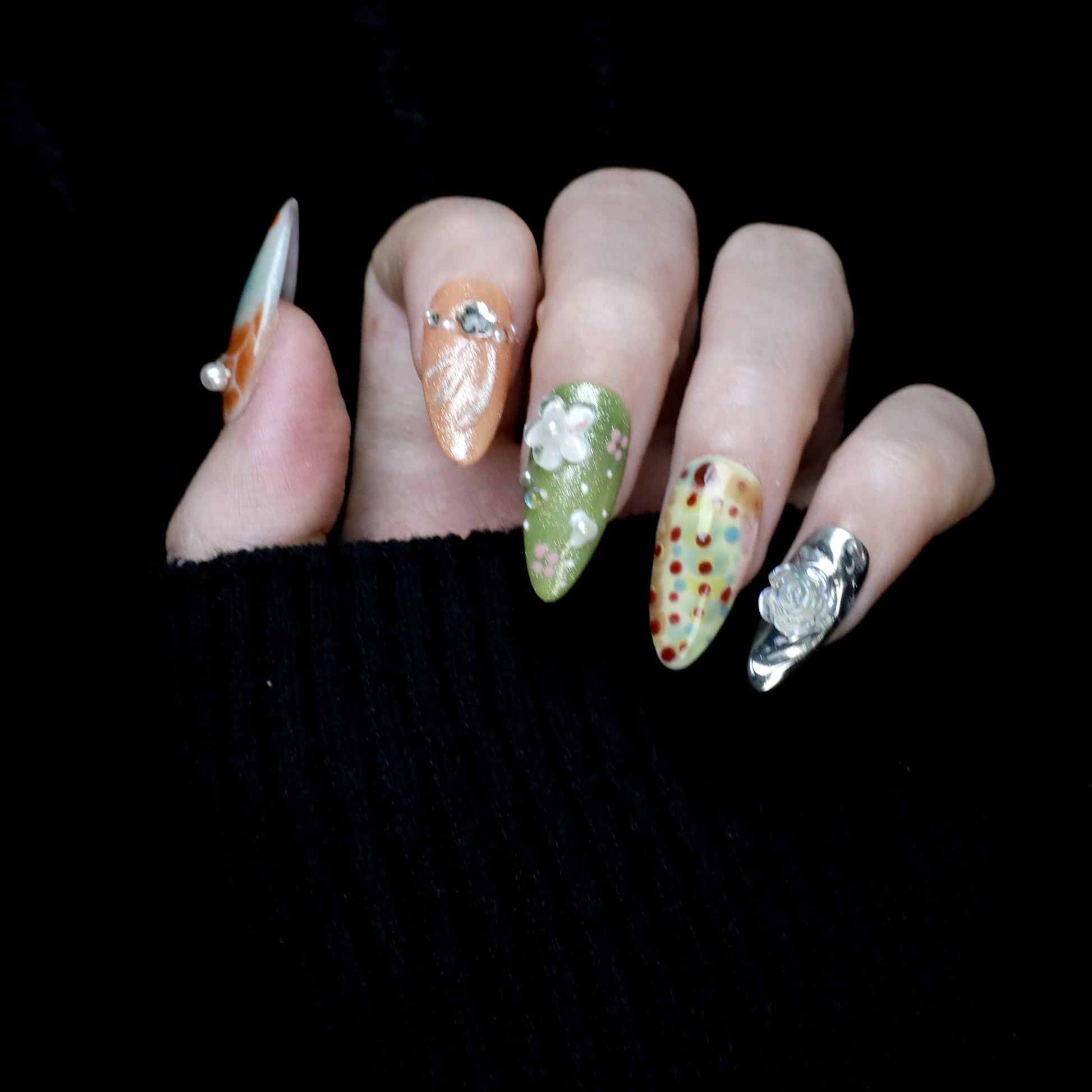 A hand showcases the Fantasy Garden Press-On Nails by We Do Nails against a black background. The nails feature designs of a koi fish with a pearl, peach with rhinestones, 3D green flowers, green with red floral dots, and metallic silver with a crystal accent.