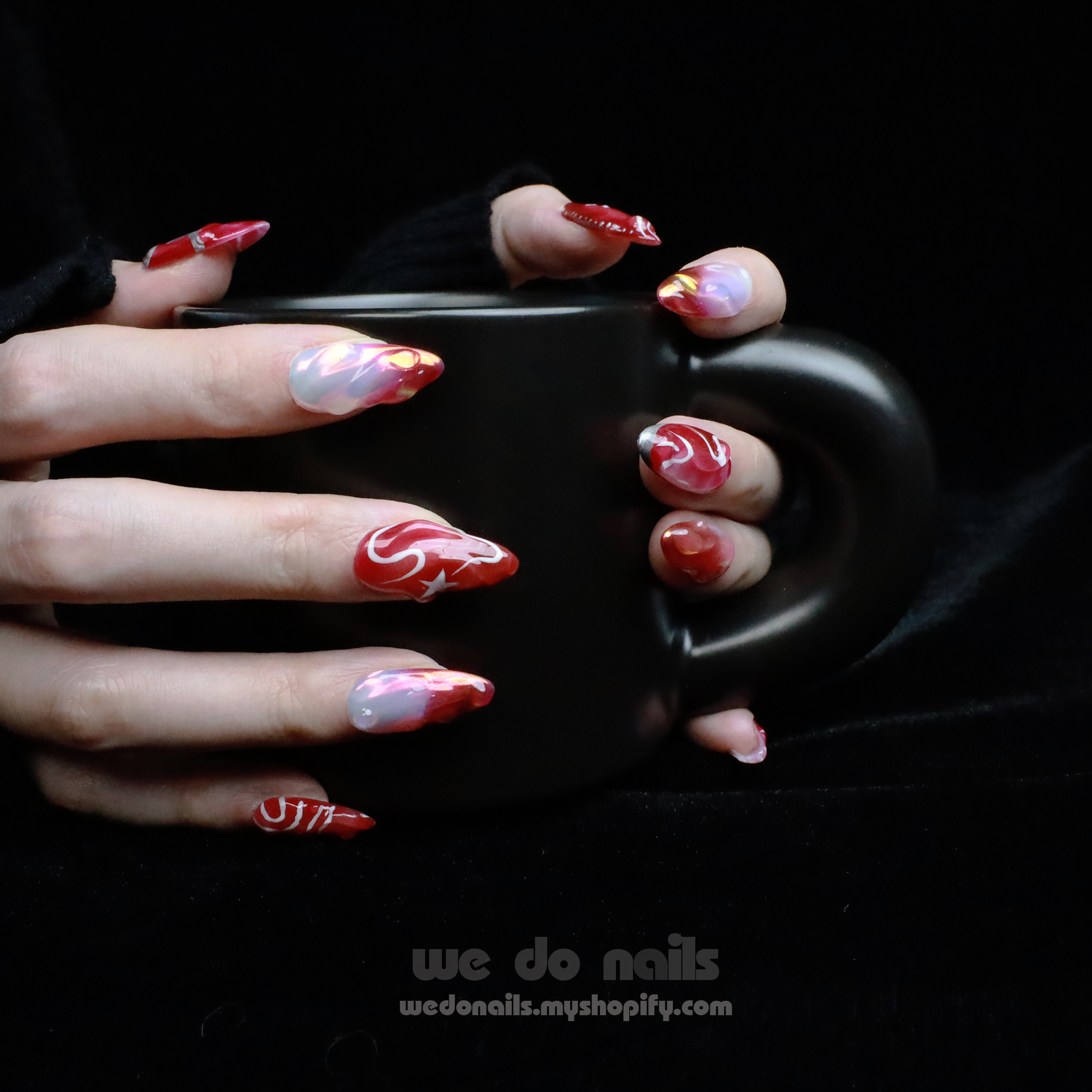A pair of hands with long, decorated red and pink nails from the Red Sky Press-On Nails by We Do Nails holds a matte black mug. The celestial glamour is evident with swirling patterns, stars, and shiny accents against a dark background—a true Starry Night Glam Nail Set masterpiece.