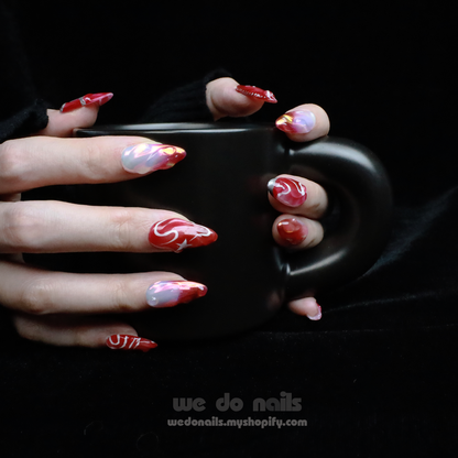 A pair of hands with long, decorated red and pink nails from the Red Sky Press-On Nails by We Do Nails holds a matte black mug. The celestial glamour is evident with swirling patterns, stars, and shiny accents against a dark background—a true Starry Night Glam Nail Set masterpiece.