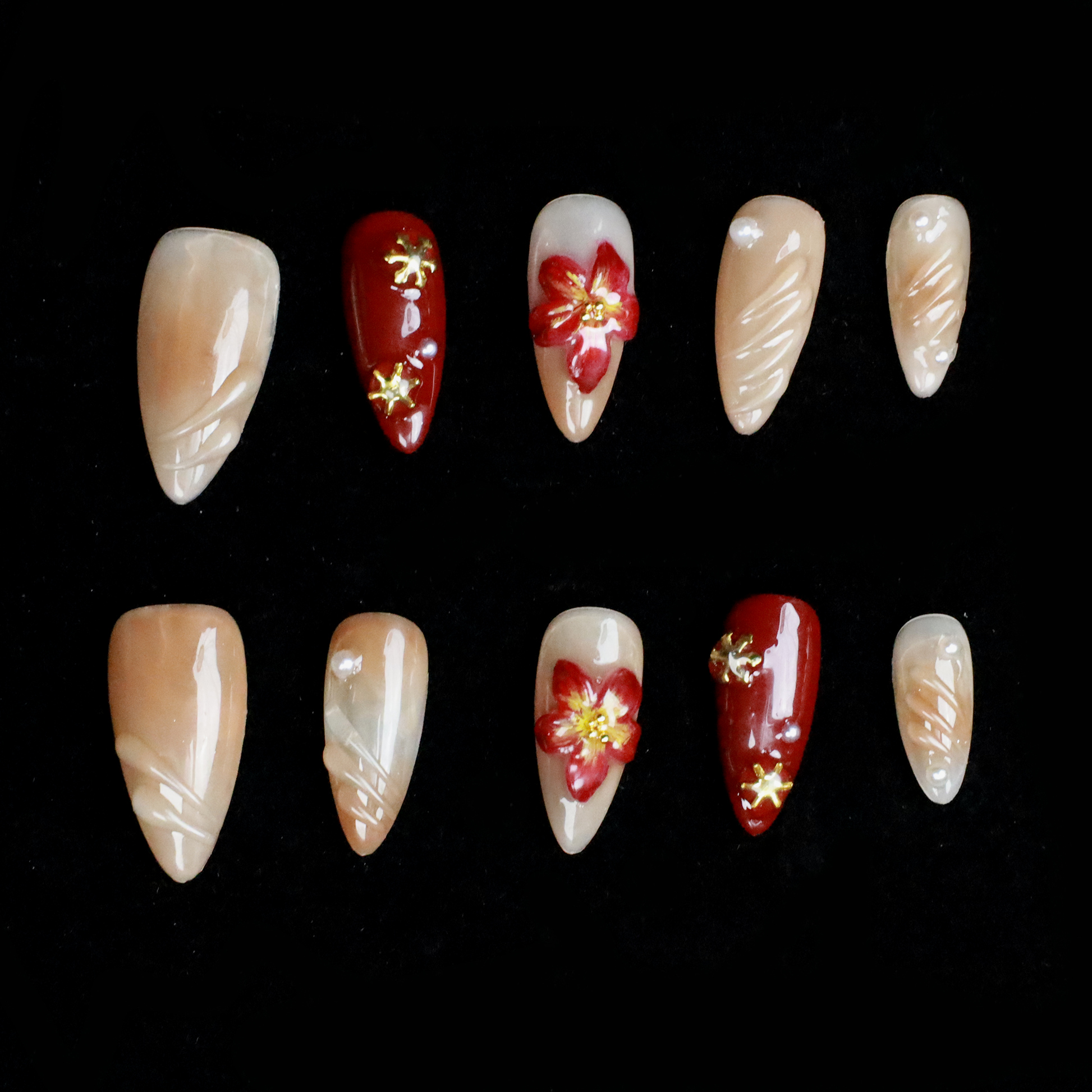 A pair of hands adorned with Elegant Floral Red and Beige Press-On Nails by We Do Nails, featuring intricate red flowers and pearl-gold accents, holds a large black mug. The elongated, almond-shaped nails boast a glossy finish with 3D floral designs against the black background.