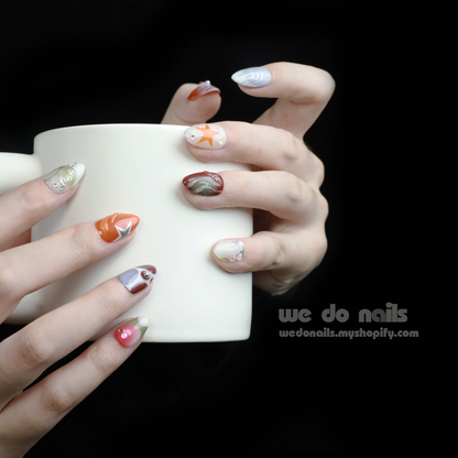 A hand displays a salon-quality manicure with Galaxy Glam Press-On Nails by We Do Nails, featuring an intricate orange and silver geometric design. The cosmic vibe shines against the dark backdrop, while SHIP NEXT DAY stands out in a pink rectangle in the upper left.