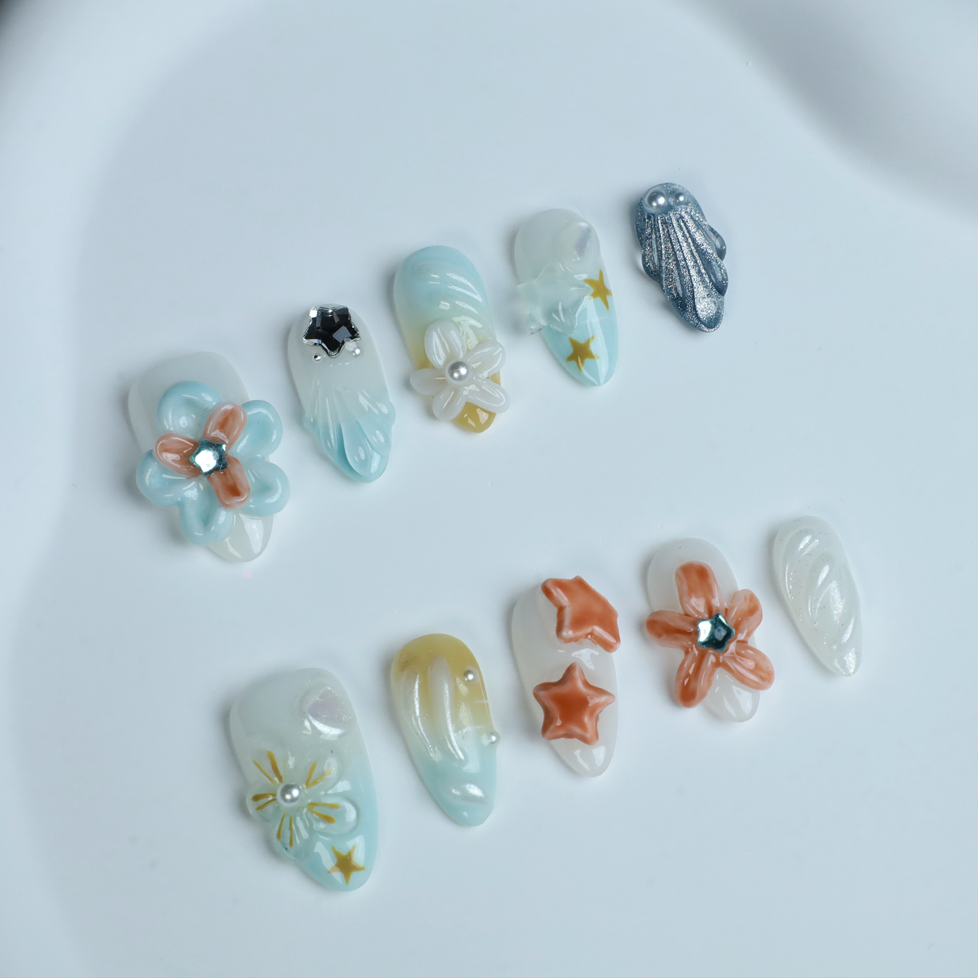 A hand with long, intricately decorated nails exhibits ethereal designs of blue and white swirls, 3D floral accents, pearls, and a metallic silver textured nail against a black background. This look perfectly captures the elegance of We Do Nails Celestial Blossom Press On Nails.