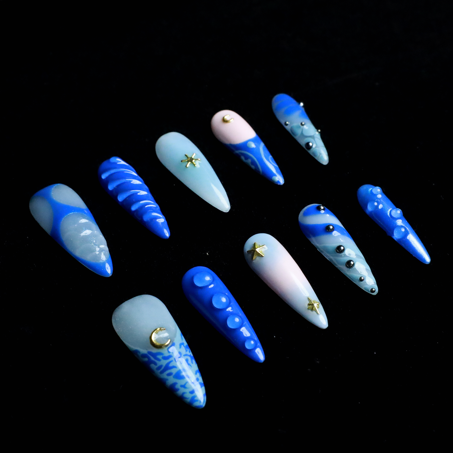 A hand featuring We Do Nails Blue Dreams Press-On Nails with elongated designs and pink hues, includes 3D metallic nail art and textured patterns like ridges and dots against a black backdrop.
