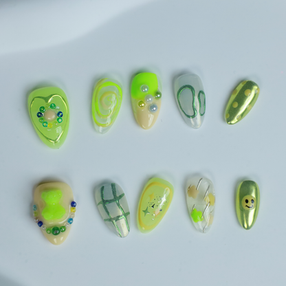A hand holding a black mug flaunts vibrant We Do Nails Neon Slime Fantasy Press-On Nails, featuring abstract lime green, yellow, and black patterns. Spirals, dots with bead accents, and 3D smiley faces create playful art against the dark backdrop.