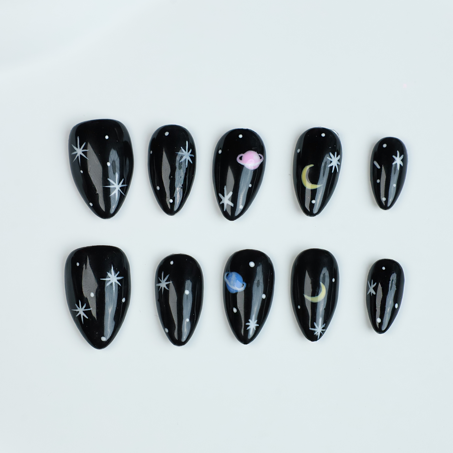 Hands decorated with Galactic Night Sky Press On Nails by We Do Nails, featuring stars, moons, and planets, hold a plain white mug against a dark backdrop.