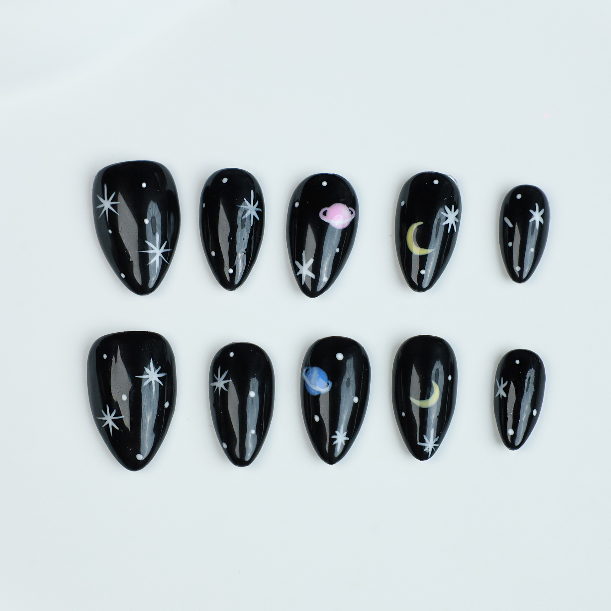 Hands decorated with Galactic Night Sky Press On Nails by We Do Nails, featuring stars, moons, and planets, hold a plain white mug against a dark backdrop.