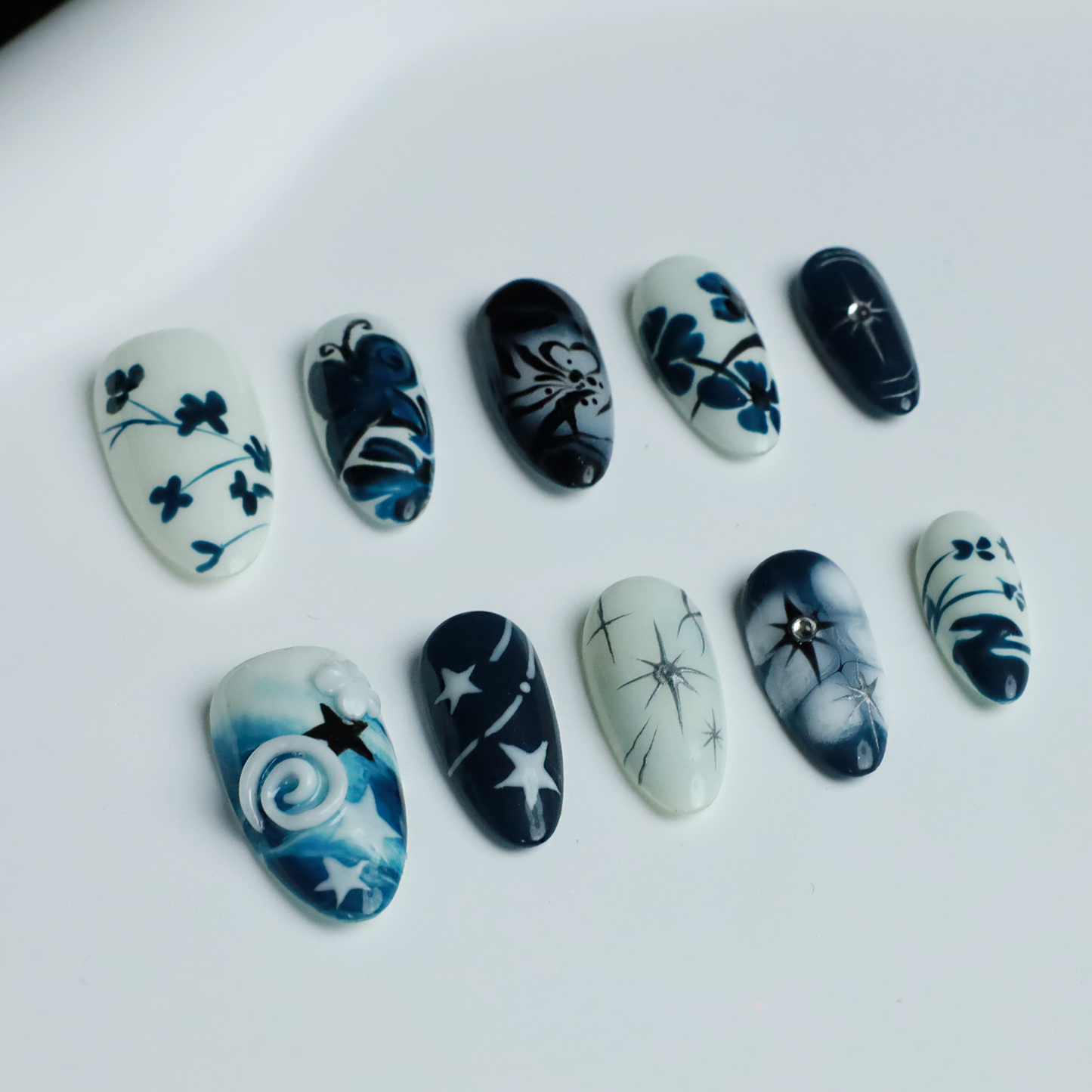 Hands adorned with We Do Nails Blue & White Porcelain Inspired Nails, showcasing long, almond-shaped designs with intricate traditional floral and abstract patterns. The nails stand out against black sleeves and a black background, reflecting the elegance of Chinese porcelain.
