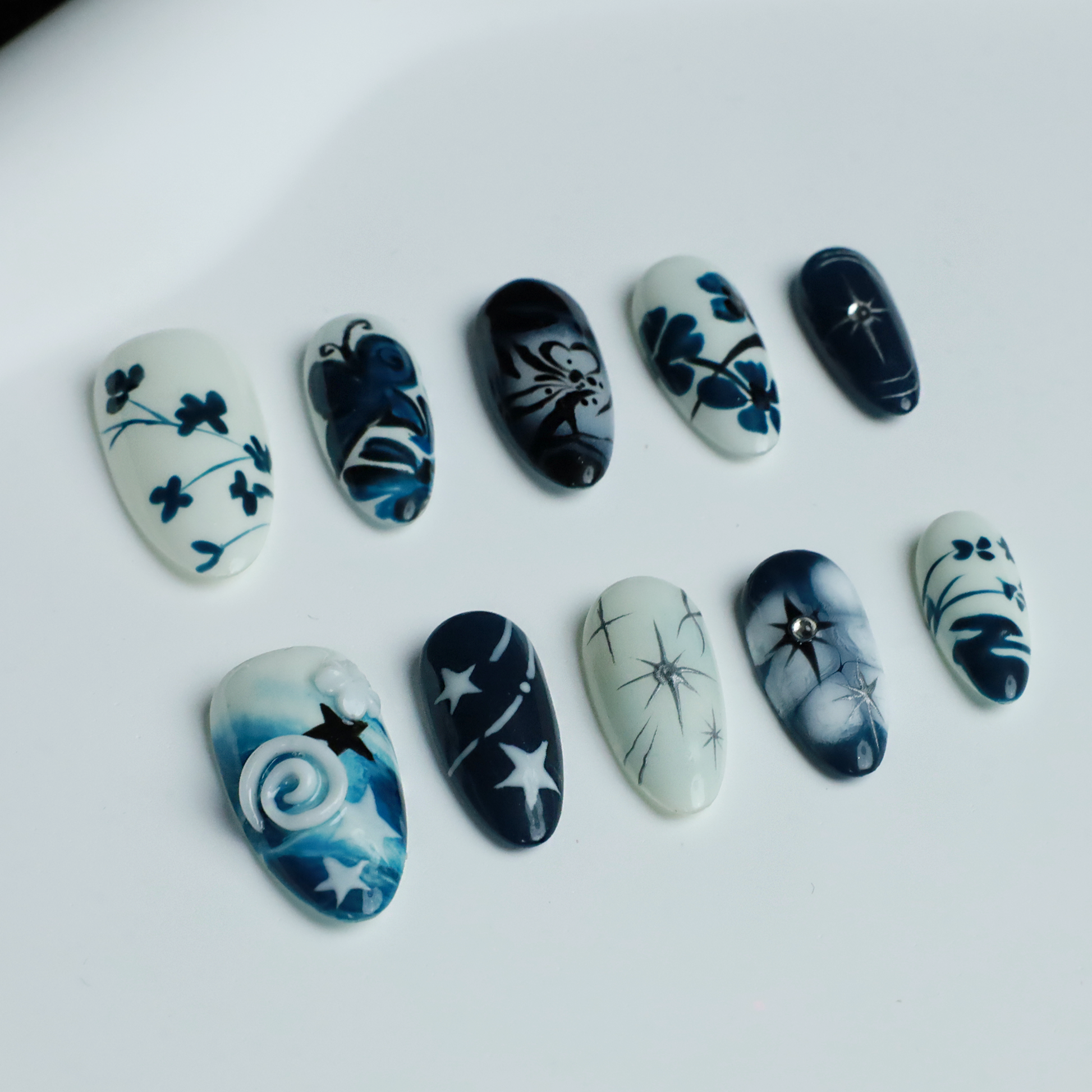 Hands adorned with We Do Nails Blue & White Porcelain Inspired Nails, showcasing long, almond-shaped designs with intricate traditional floral and abstract patterns. The nails stand out against black sleeves and a black background, reflecting the elegance of Chinese porcelain.