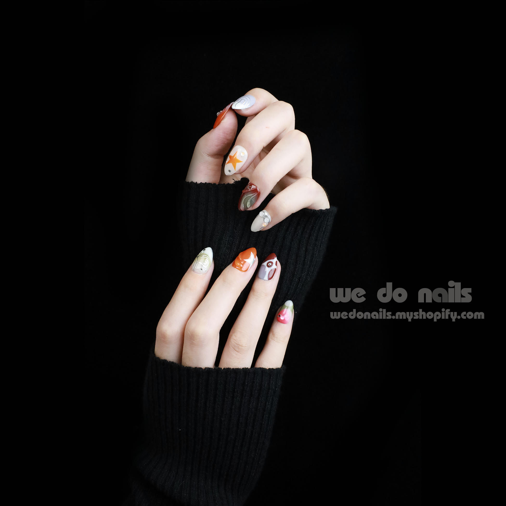 A hand displays a salon-quality manicure with Galaxy Glam Press-On Nails by We Do Nails, featuring an intricate orange and silver geometric design. The cosmic vibe shines against the dark backdrop, while SHIP NEXT DAY stands out in a pink rectangle in the upper left.