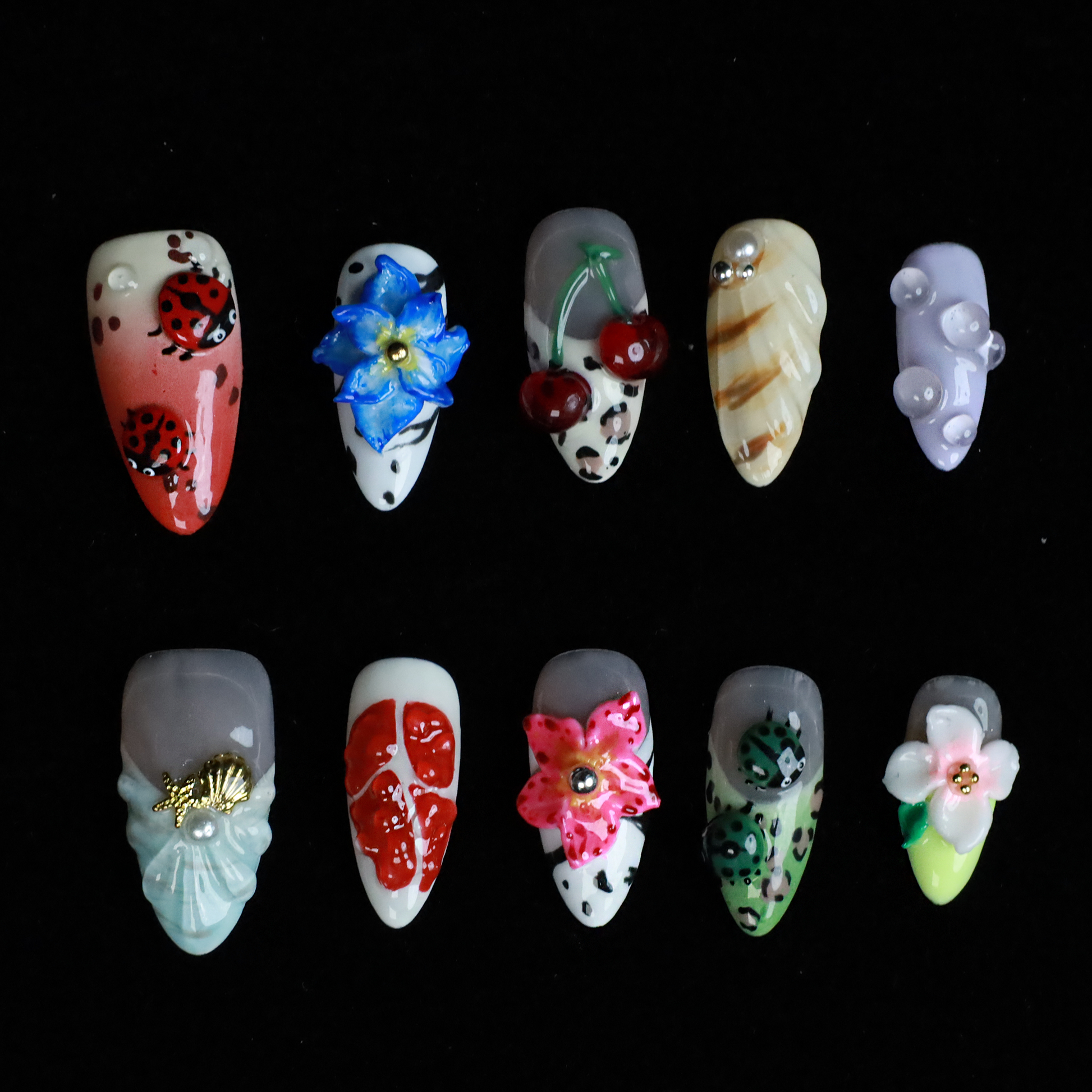The Garden Blooms Press-On Nails by We Do Nails feature a whimsical garden theme with long, decorative nails, including a blue flower, cherries, red and white patterns, pearls, and stripes. Set against a black background, the hand is partially covered by a black sleeve.