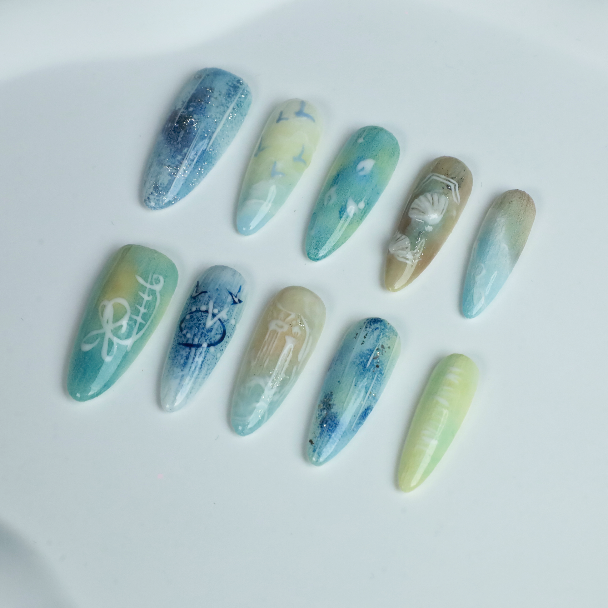 A hand flaunts the elegant Ocean-Inspired Press-On Nails by We Do Nails, featuring long, pointed shapes with ocean-themed designs. The set showcases ocean breeze colors, seashell art, and wave patterns against a dark background to highlight its intricate beauty.