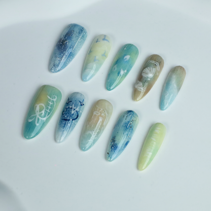 A hand flaunts the elegant Ocean-Inspired Press-On Nails by We Do Nails, featuring long, pointed shapes with ocean-themed designs. The set showcases ocean breeze colors, seashell art, and wave patterns against a dark background to highlight its intricate beauty.