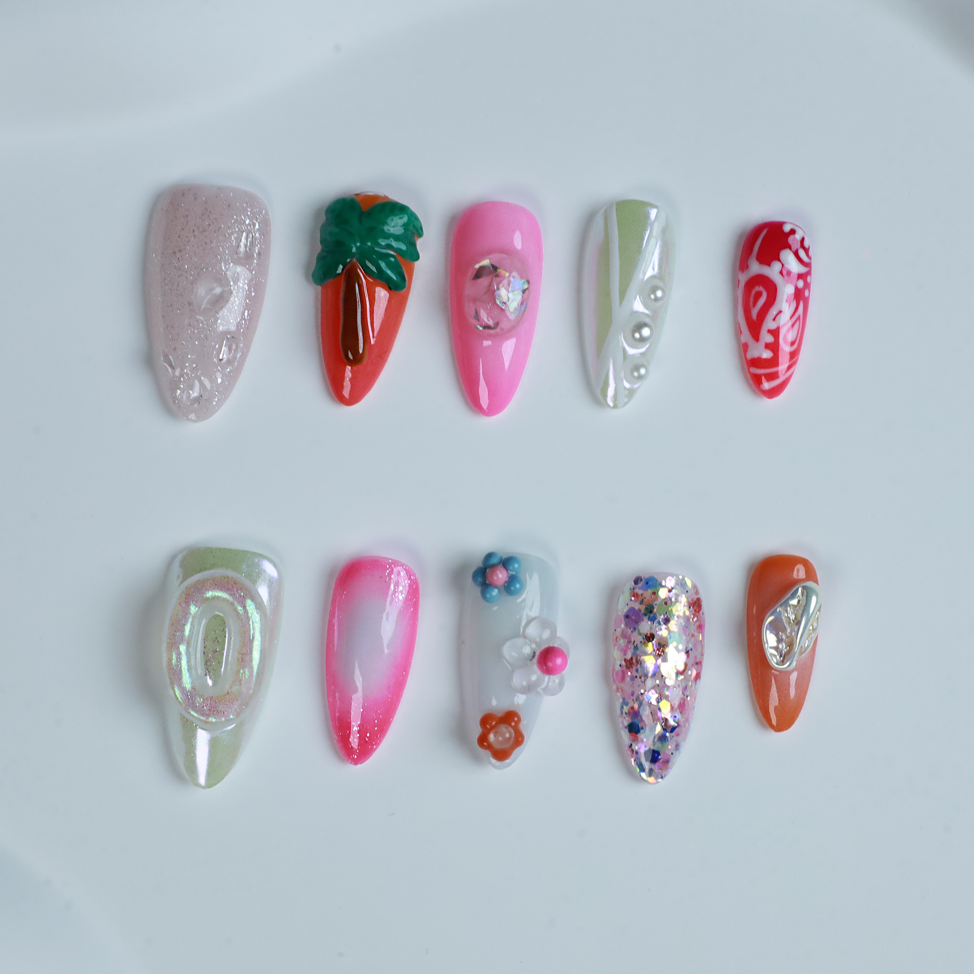 A hand features the Summer Island Press-On Nails by We Do Nails: glossy pearl-adorned, carrot-themed orange with neon, pastel pink in floral art, translucent with pearls, and red nails highlighted by an abstract white design.