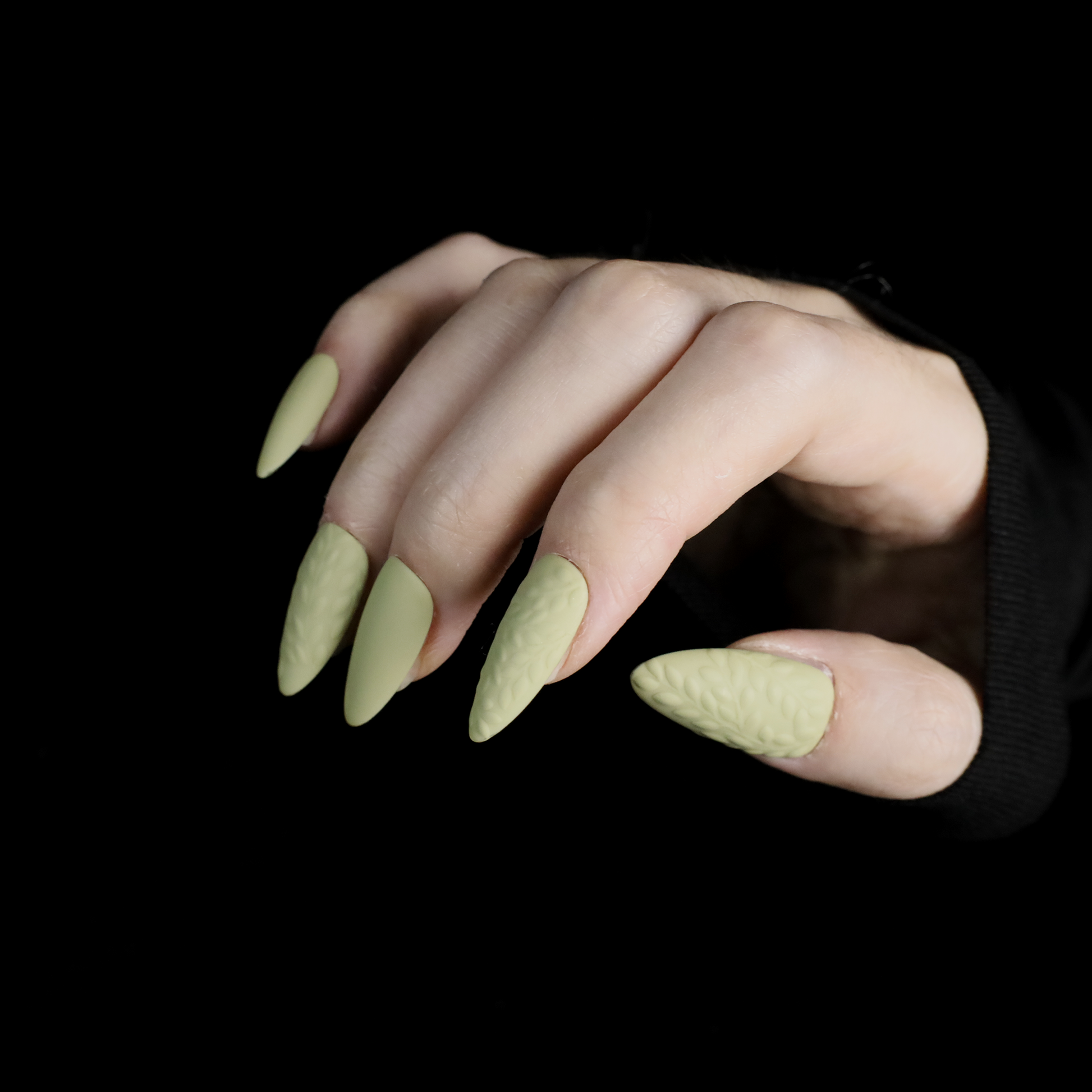 A person displays their crossed hands against a black background, showcasing We Do Nails Earthy Olive Green Press-On Nails. The textured olive design is elegantly highlighted by the sleeves of their black garment.