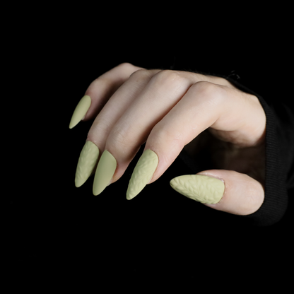 A person displays their crossed hands against a black background, showcasing We Do Nails Earthy Olive Green Press-On Nails. The textured olive design is elegantly highlighted by the sleeves of their black garment.