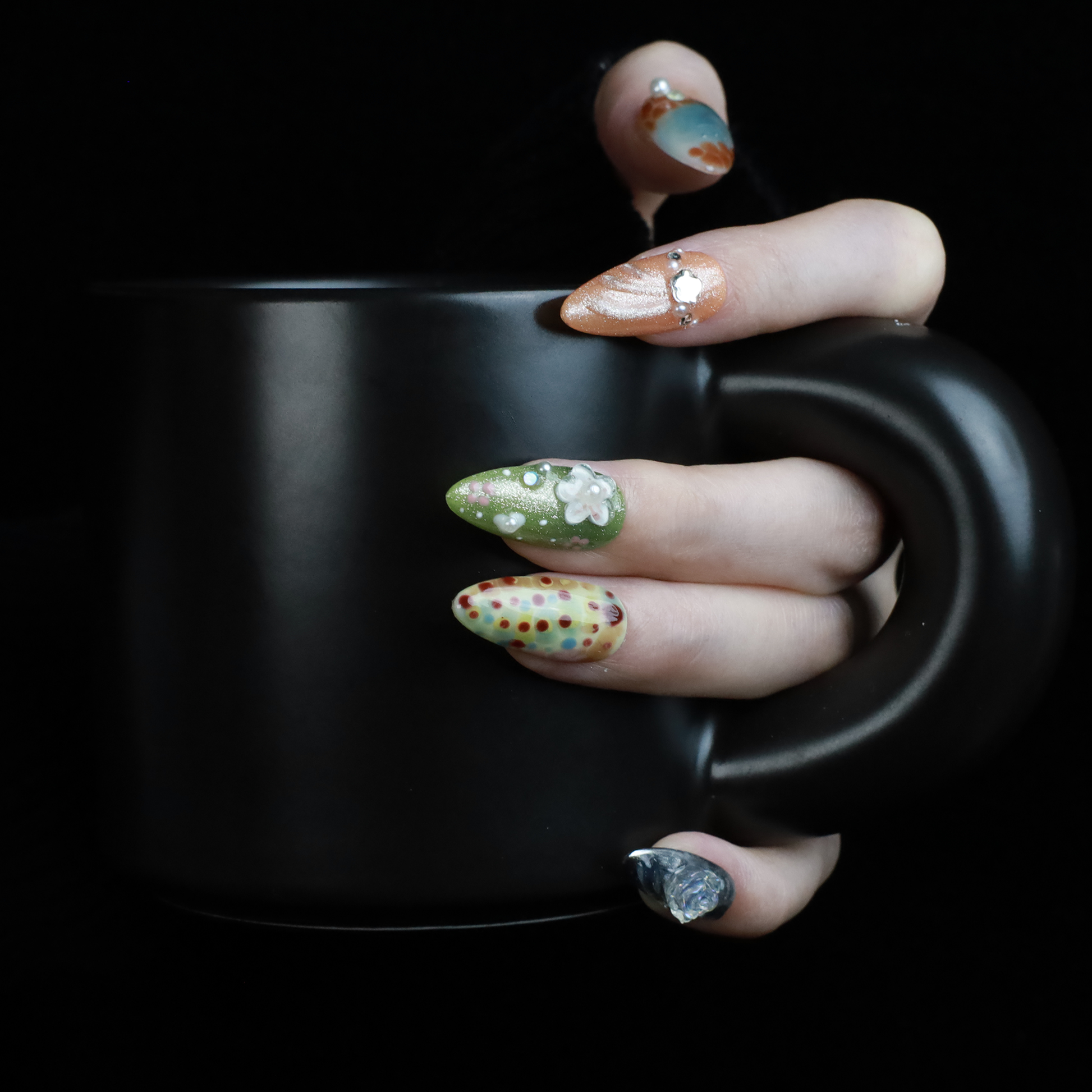 A hand showcases the Fantasy Garden Press-On Nails by We Do Nails against a black background. The nails feature designs of a koi fish with a pearl, peach with rhinestones, 3D green flowers, green with red floral dots, and metallic silver with a crystal accent.