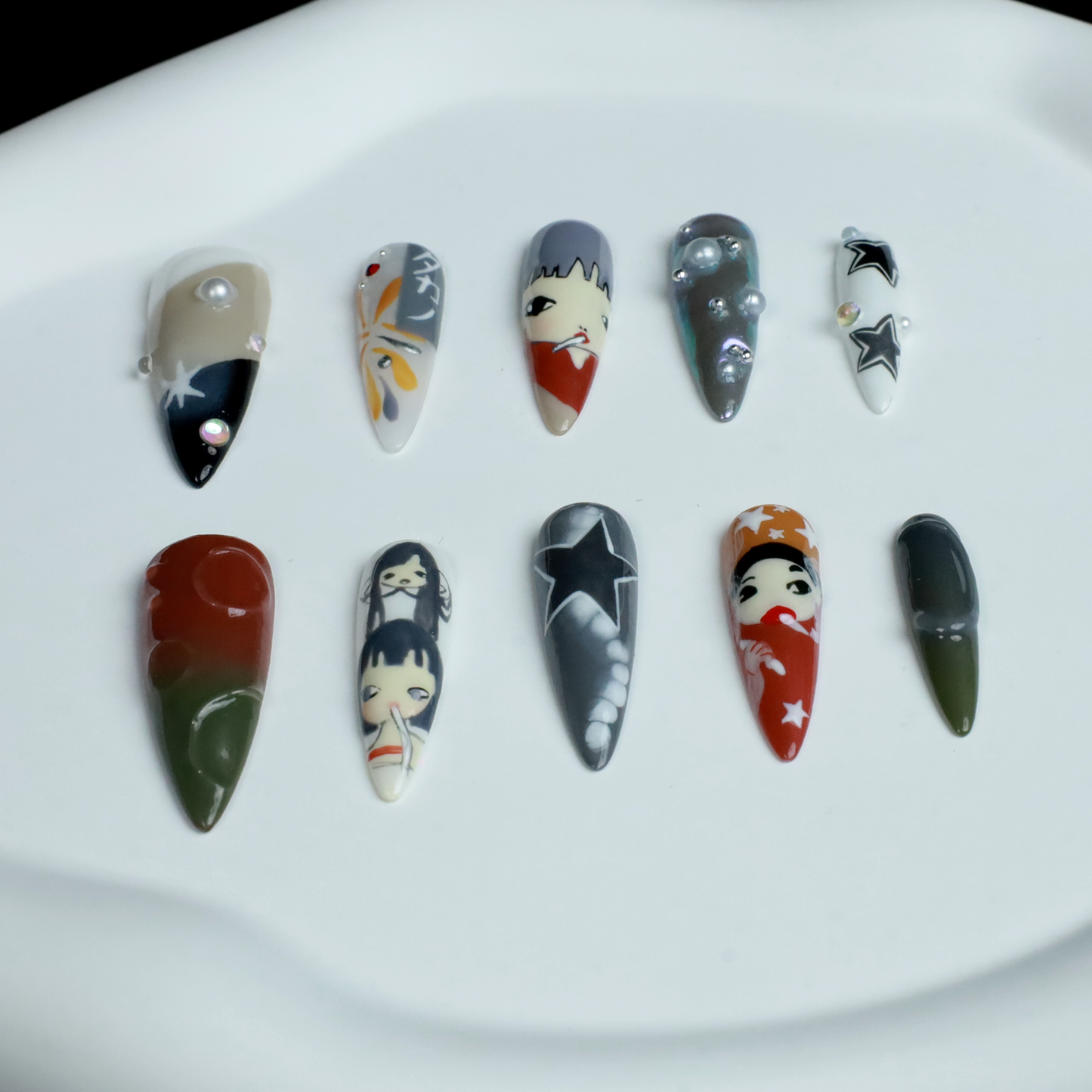 A hand with long, pointed Japanese-Inspired Artistic Press-On Nails by We Do Nails showcases camo patterns in green, red, and gray. Combining kawaii aesthetic and artistic motifs that suggest anime nail art, the hand is partially covered by a black sleeve.