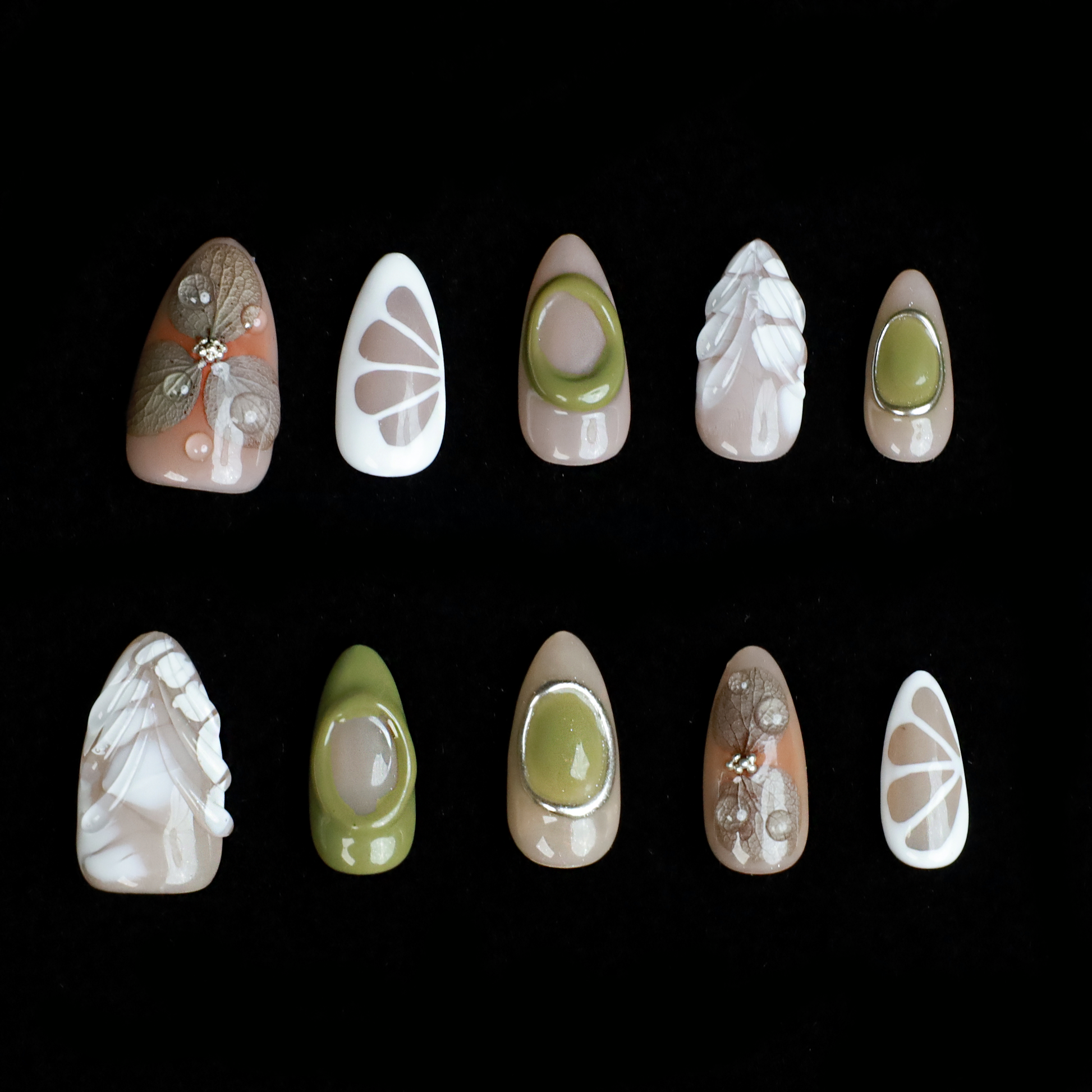 A hand in a black sleeve displays the Leaf and Organic design Press-On Nails by We Do Nails, featuring long acrylic nails with white abstract patterns, green accents, and clear 3D embellishments that create a nature-inspired look against the dark background.
