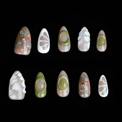 A hand in a black sleeve displays the Leaf and Organic design Press-On Nails by We Do Nails, featuring long acrylic nails with white abstract patterns, green accents, and clear 3D embellishments that create a nature-inspired look against the dark background.