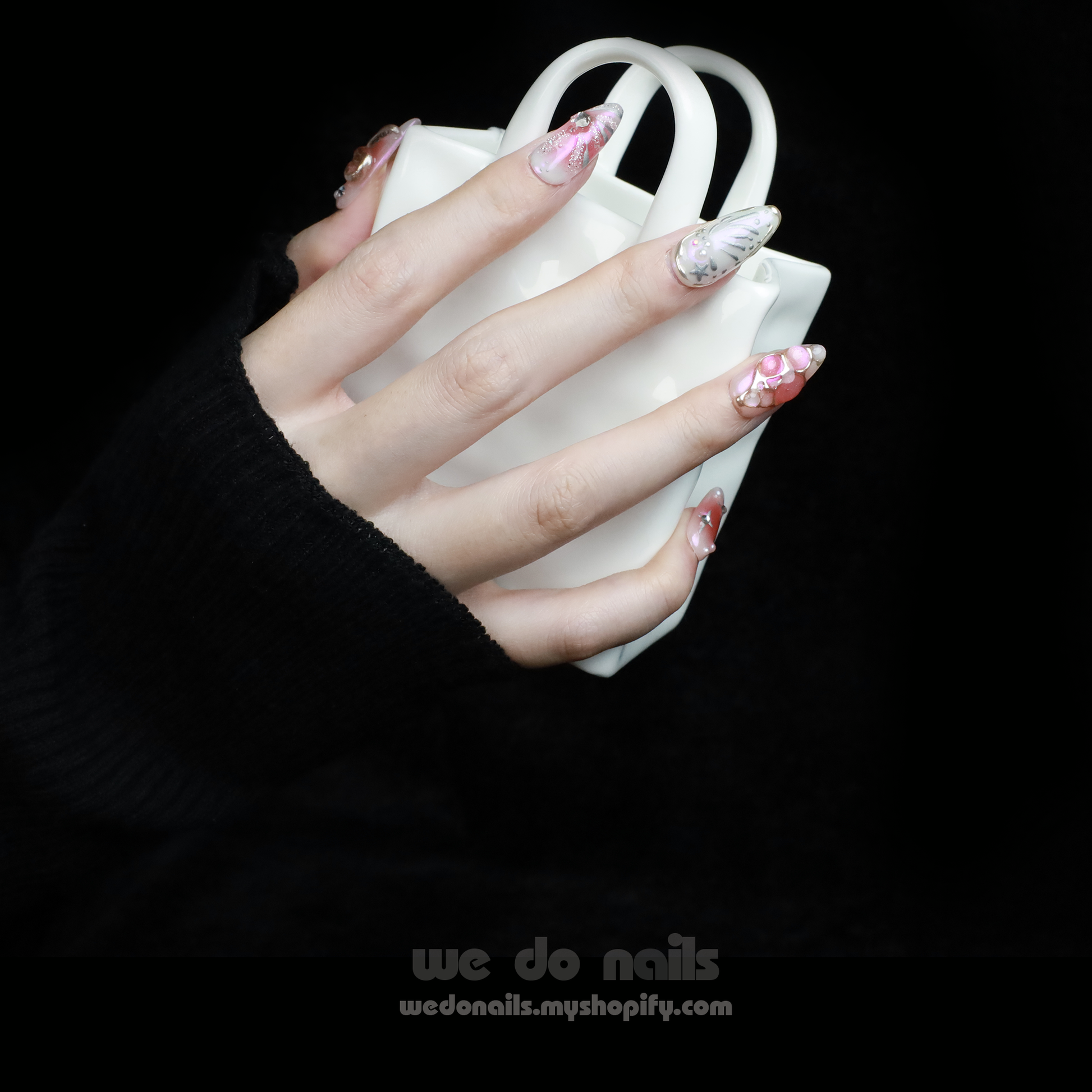A person with intricately designed Blushing Charm Press-On Nails by We Do Nails holds a small, white ceramic handbag. The nails display soft pinks and abstract patterns in pink, white, and gold, accented with shimmering details and decorative embellishments.