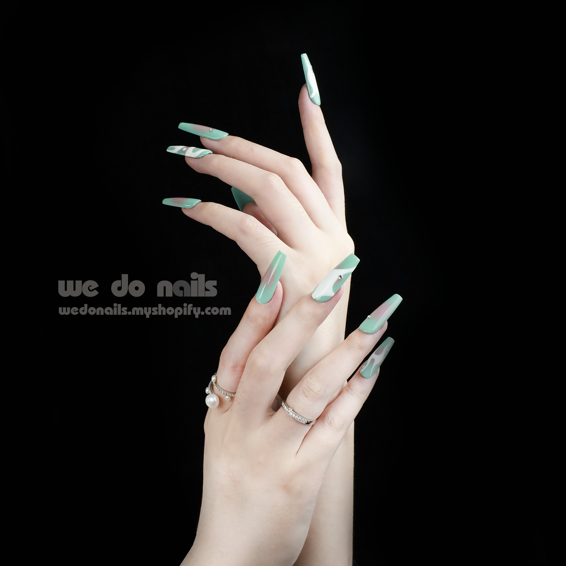 Two hands featuring We Do Nails Spilled Milk Press-On Nails, adorned with a modern, green geometric pattern, are elegantly poised against a black background. The fingers wear multiple rings and showcase the artistic nail design with a small text logo visible.