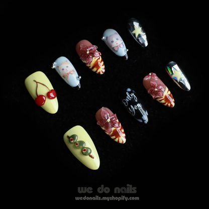 A hand adorned with We Do Nails Extravaganza Press-On Nails, showcasing unique patterns and colors, holds a small white handbag. The dazzling metallics gleam against the black background, highlighting the bold nail art and bag.