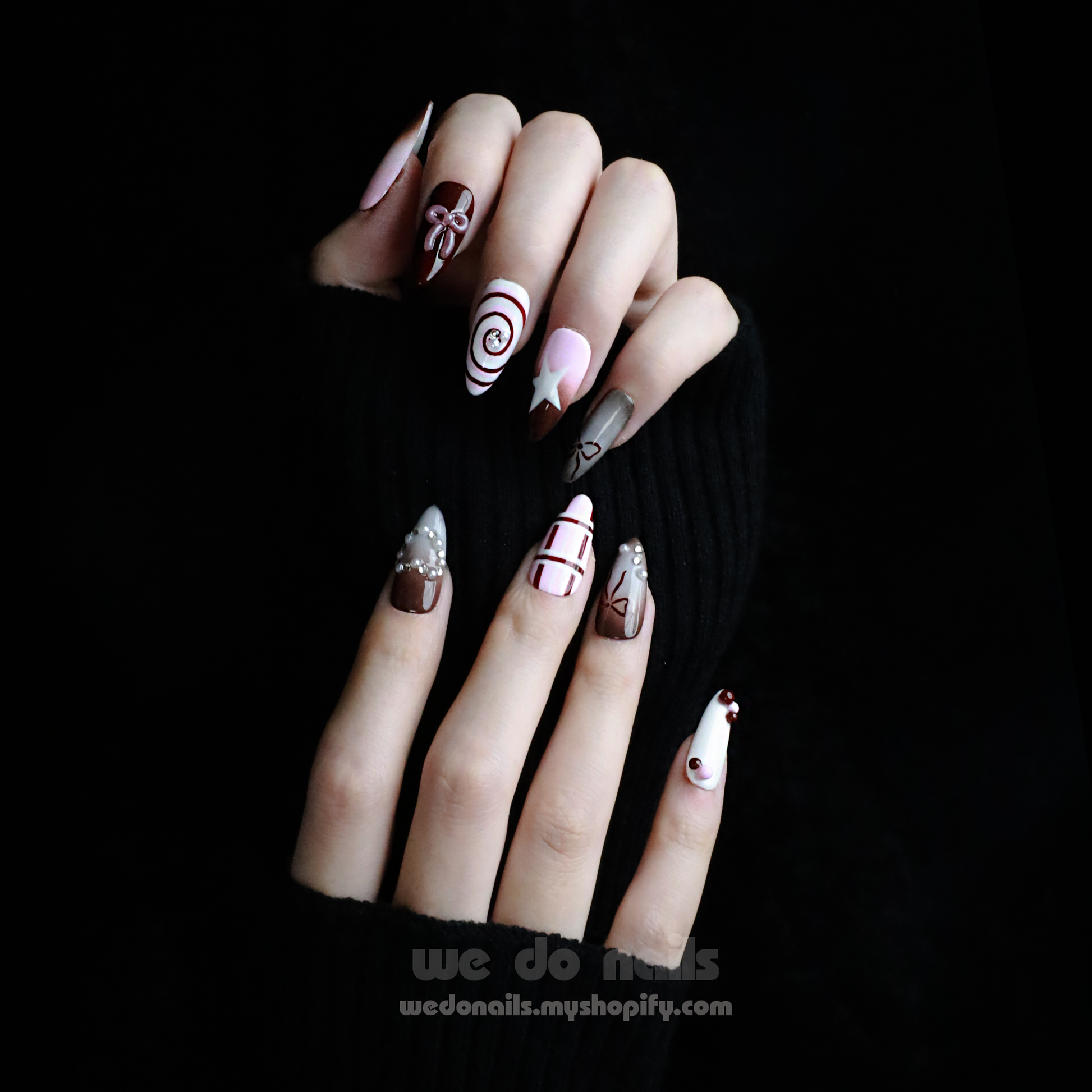 A hand with enchanting nails holds a small white handbag. The Charming Cocoa Delight Press-On Nails from We Do Nails showcase intricate patterns, featuring stripes and dotted embellishments in white, brown, and red shades, all set against a black background that enhances their allure.