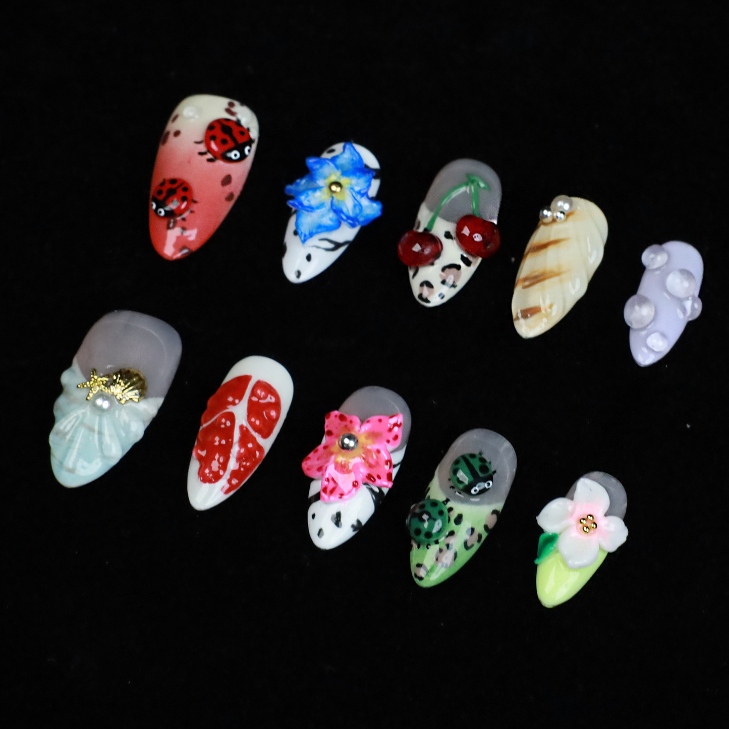 The Garden Blooms Press-On Nails by We Do Nails feature a whimsical garden theme with long, decorative nails, including a blue flower, cherries, red and white patterns, pearls, and stripes. Set against a black background, the hand is partially covered by a black sleeve.