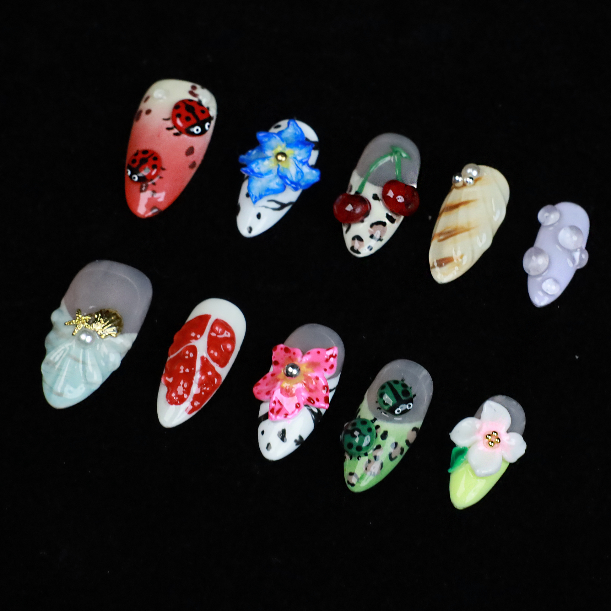 The Garden Blooms Press-On Nails by We Do Nails feature a whimsical garden theme with long, decorative nails, including a blue flower, cherries, red and white patterns, pearls, and stripes. Set against a black background, the hand is partially covered by a black sleeve.