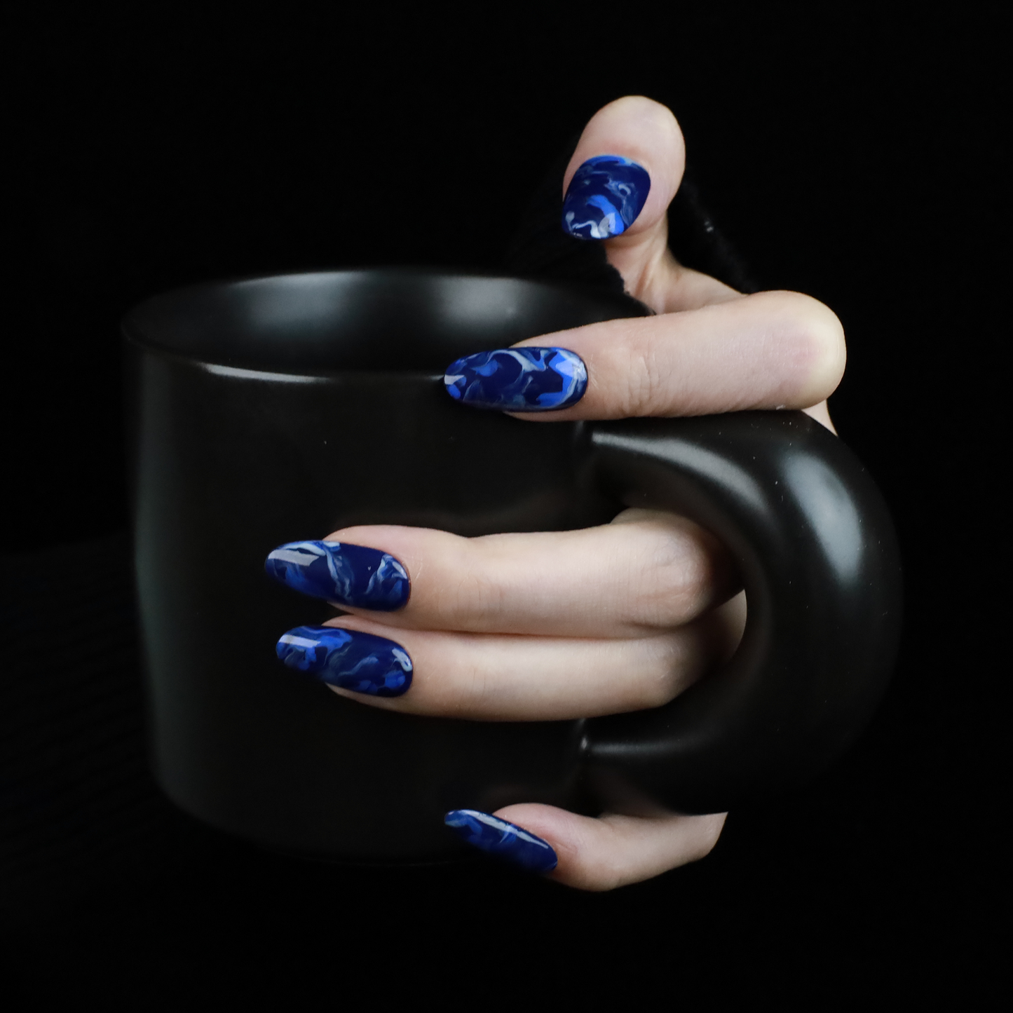 Hands display the Elegant Blue Marble Press-On Nails from We Do Nails, featuring glossy long oval shapes with a blue marble effect. A black ribbed sweater against a black background highlights these striking nails.