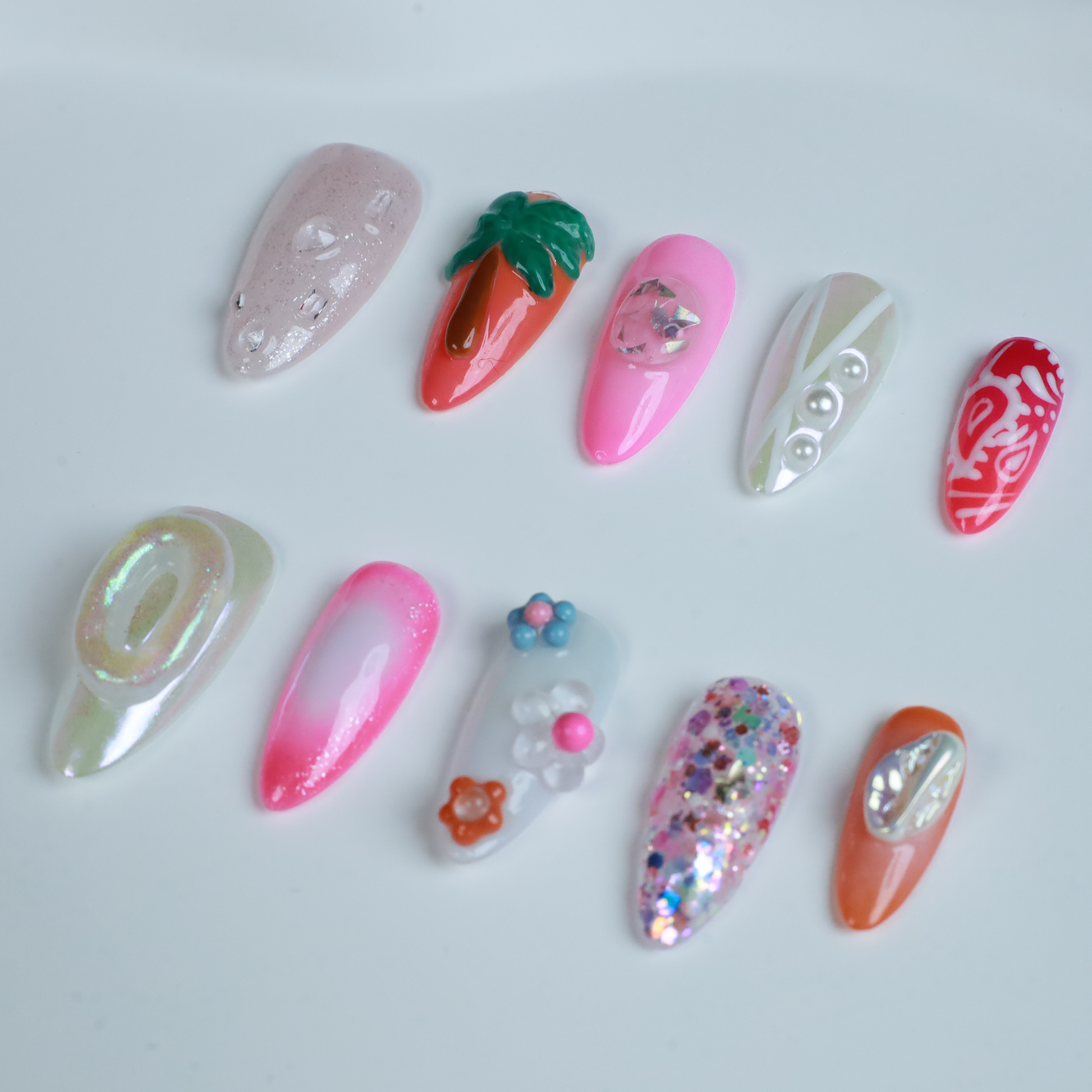 A hand features the Summer Island Press-On Nails by We Do Nails: glossy pearl-adorned, carrot-themed orange with neon, pastel pink in floral art, translucent with pearls, and red nails highlighted by an abstract white design.